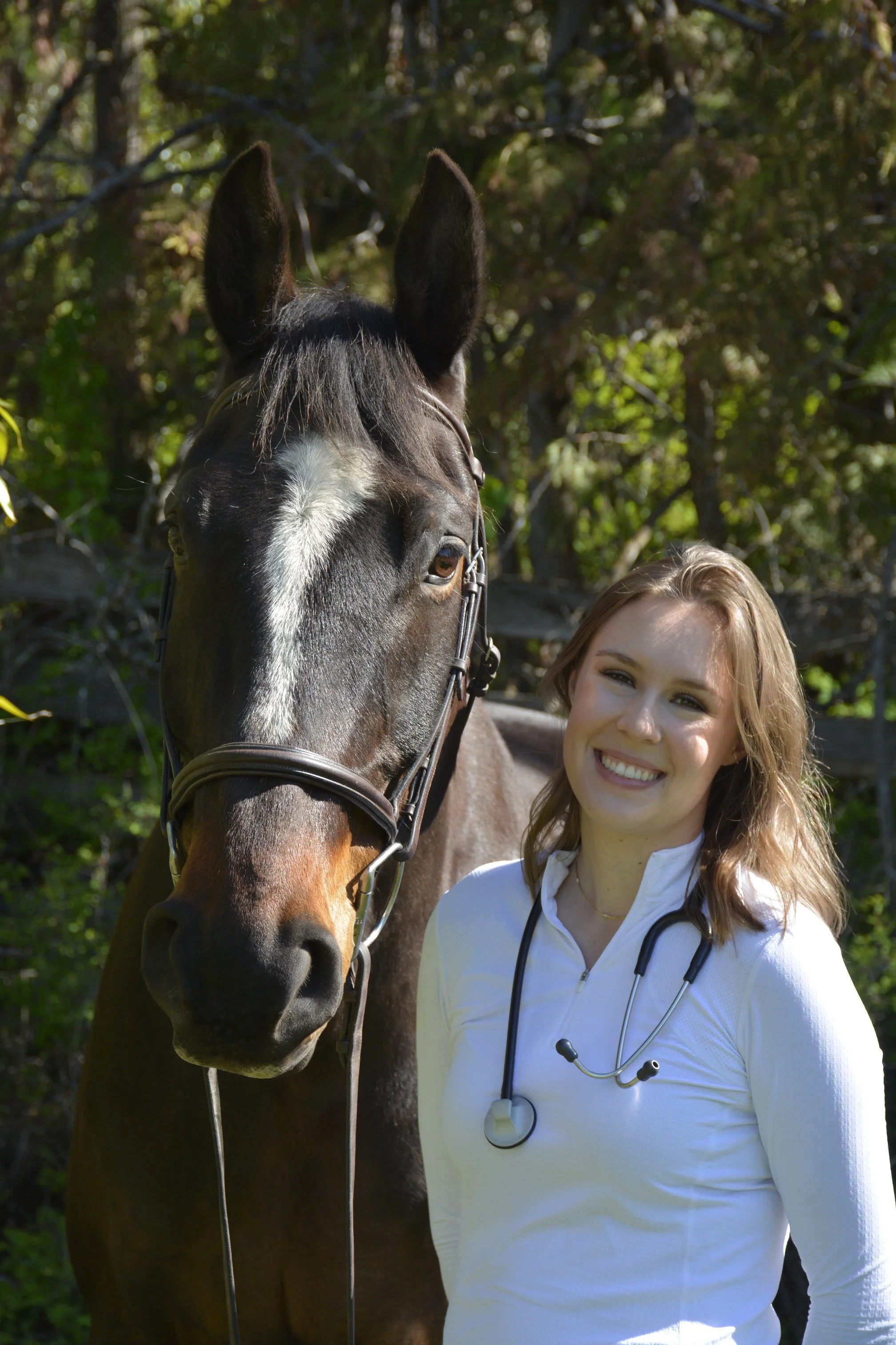 A smiling woman with a stethoscope around her neck standing next to a horse in an outdoor, wooded area.