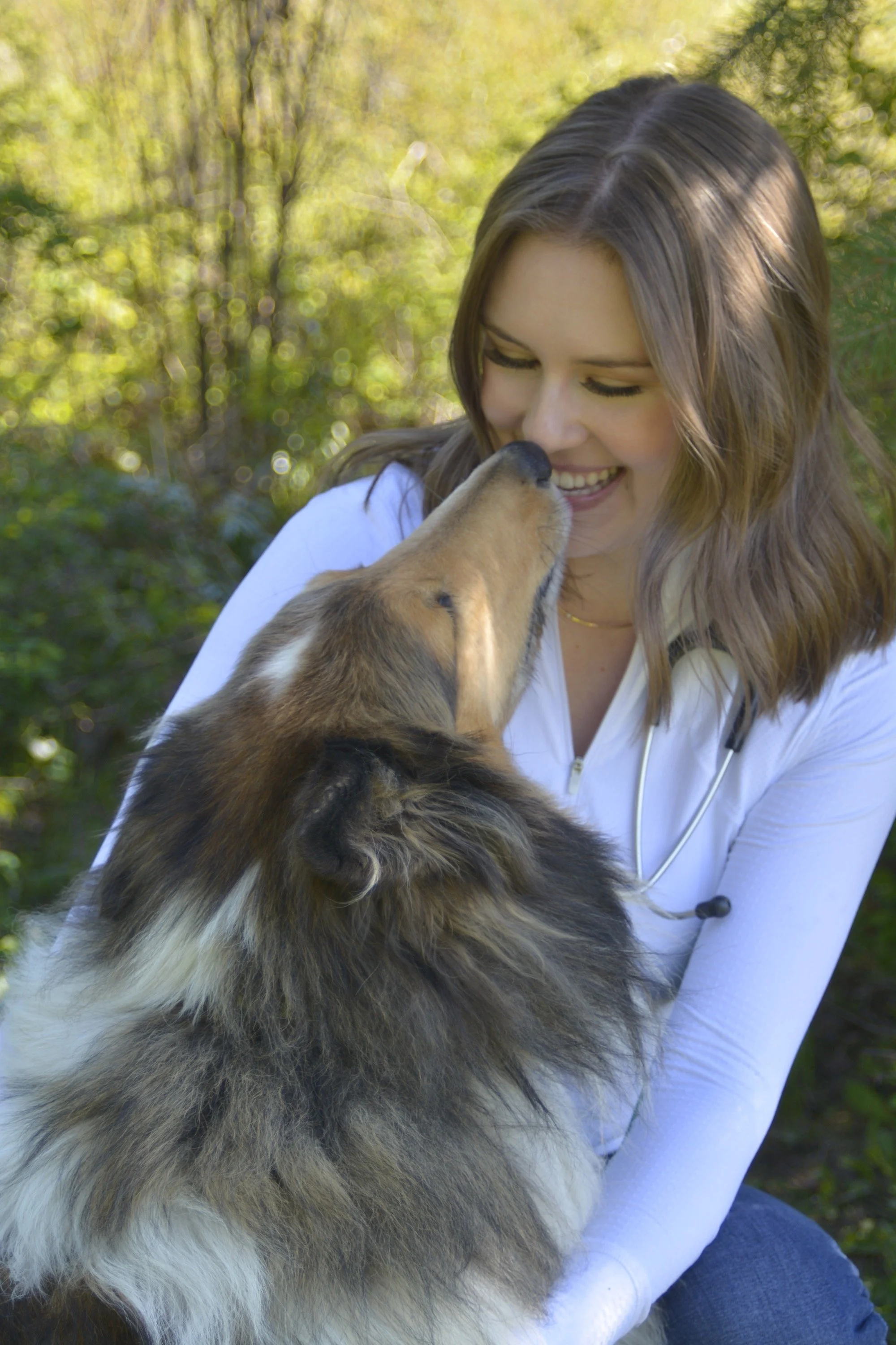 A woman with long brown hair smiling while her nose is being licked by a dog outdoors, with a background of green trees.