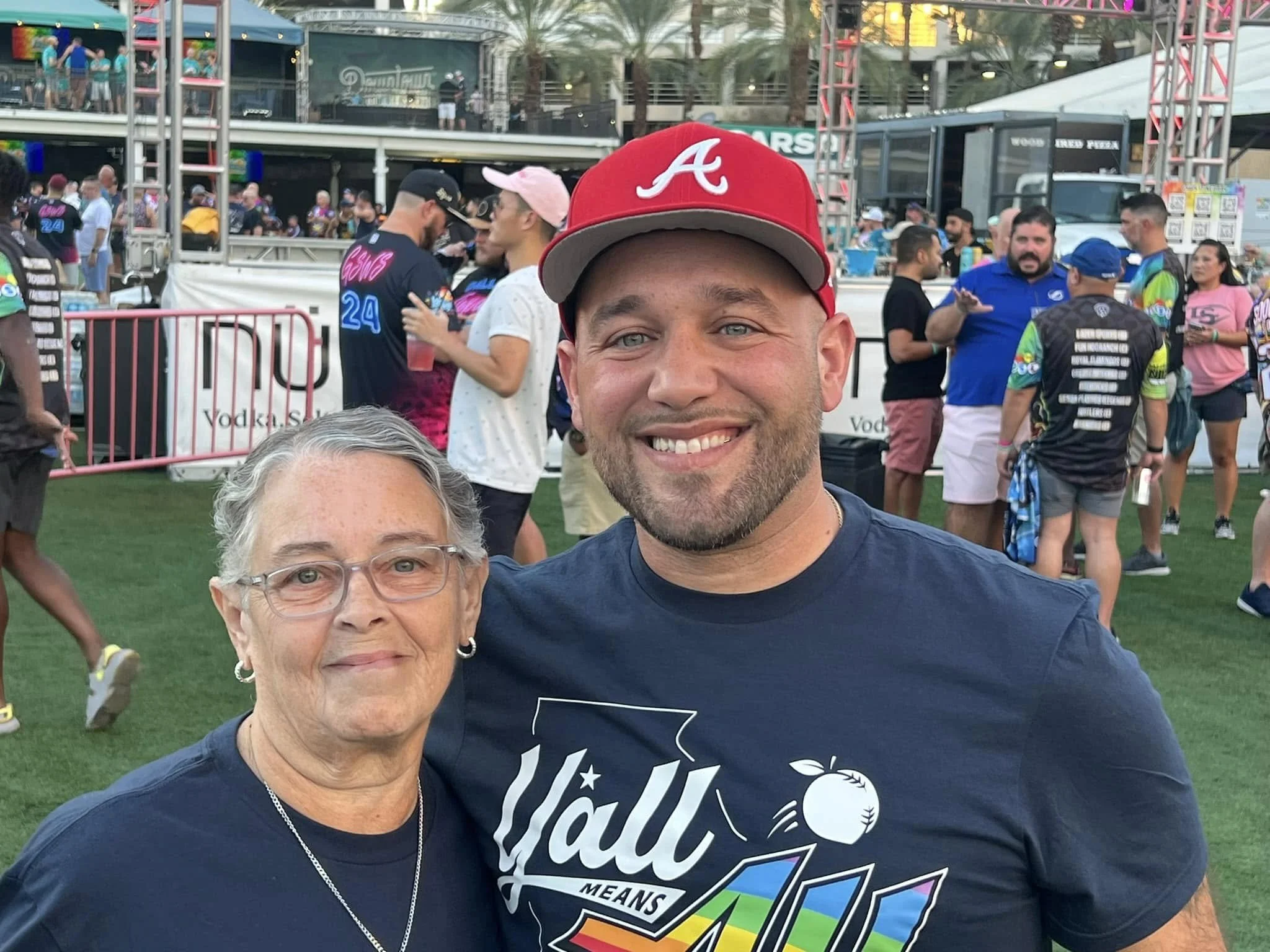 A smiling young man wearing a red baseball cap with the 'A' logo, and a woman with glasses in the foreground, at an outdoor event with a crowd of people, tents, and booths in the background.