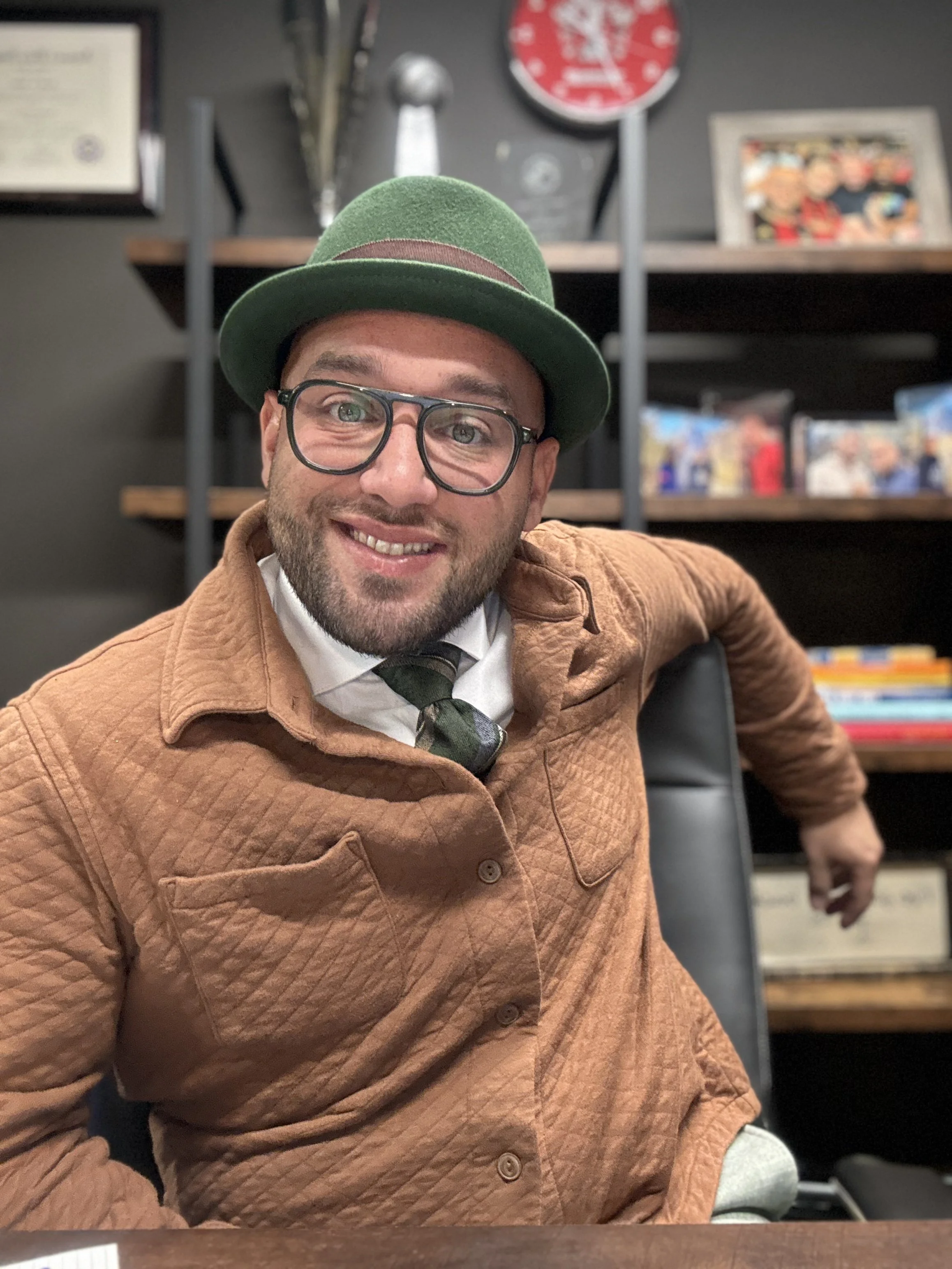 A man with glasses and a beard wearing a green felt fedora hat, a brown quilted jacket, white shirt, and a tie, sitting in an indoor space with shelves containing books and framed pictures in the background.