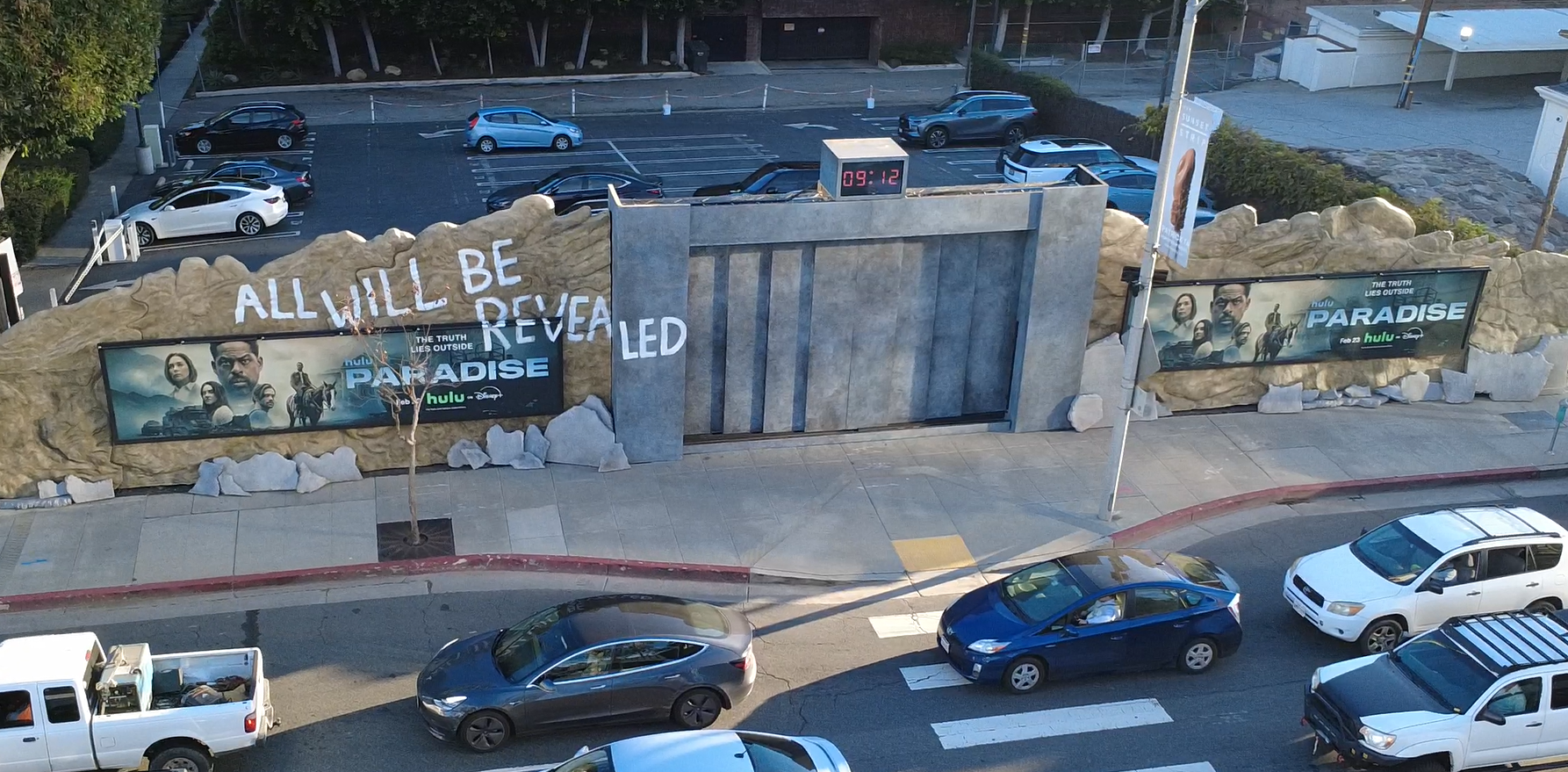 A parking lot with cars in front of a rocky wall with the words 'ALL WILL BE REVEALED' painted on it. There are two digital clocks showing the time 09:12, and electronic billboards advertising the show 'Paradise' on Hulu. The scene appears to be take