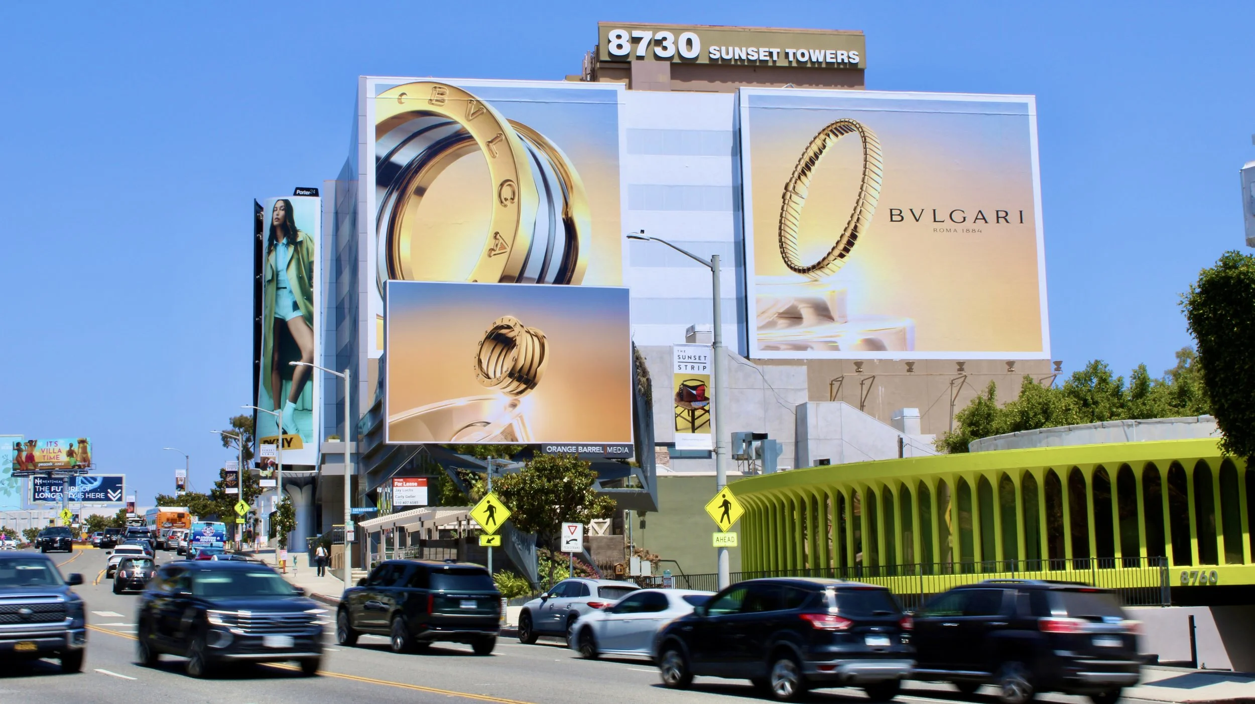 Street scene with large billboards advertising jewelry brands Bougalri and rings at Sunset Towers, blue sky, cars on the road, crosswalk signs, and a pedestrian bridge with a green railing.