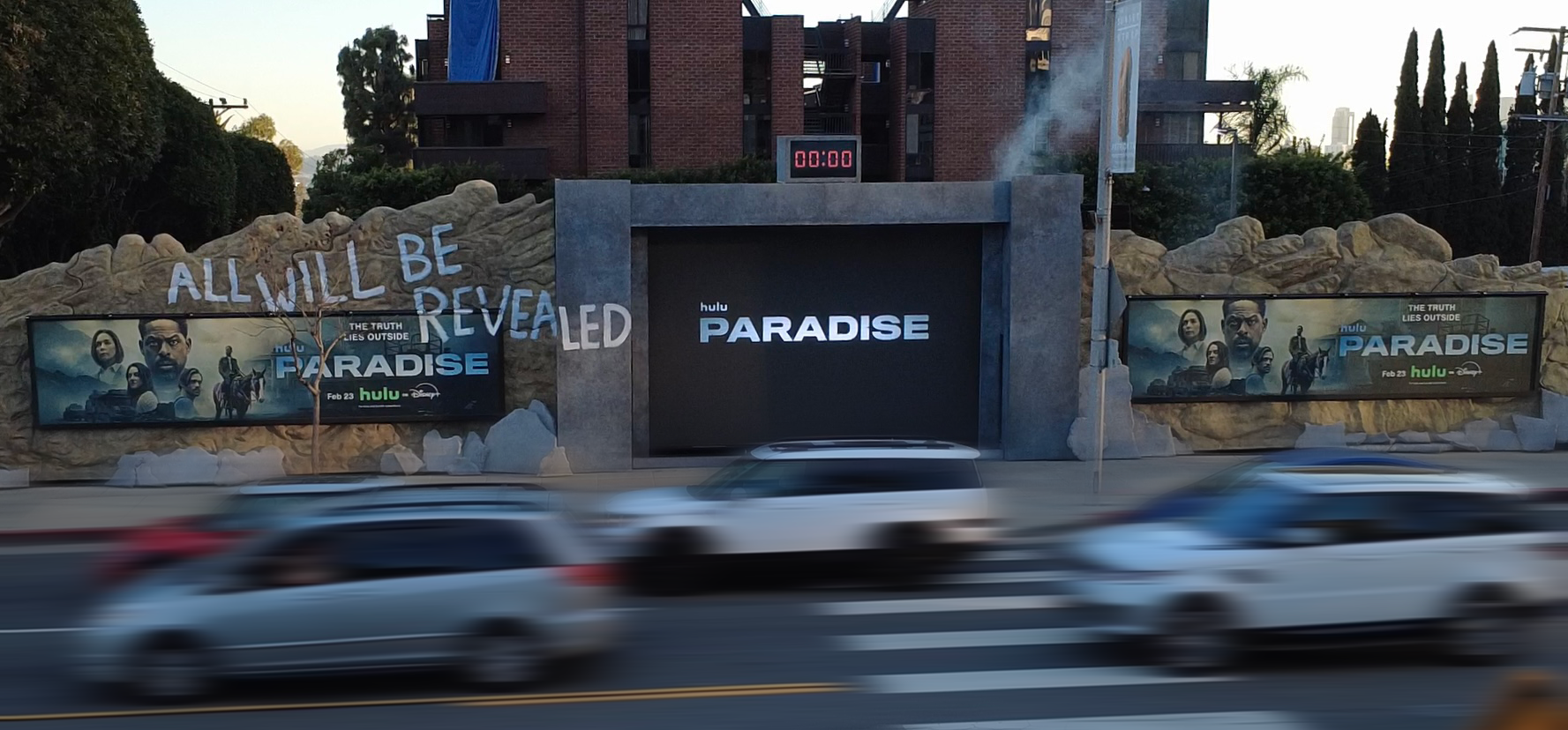 Billboard and digital sign advertising the Hulu series 'Paradise,' with additional promotional posters on a rock wall, and vehicles passing by on a road in the foreground.