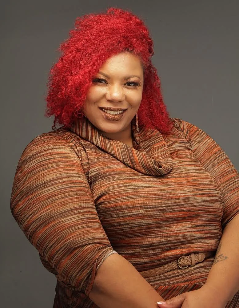 A woman with vibrant red curly hair smiling, wearing a multicolored striped dress with a wide collar and a belt, posed against a plain gray background.