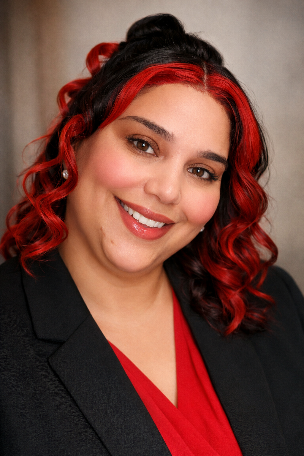 A woman with brown eyes and curly hair with red highlights, wearing a black blazer and red top, smiling at the camera.