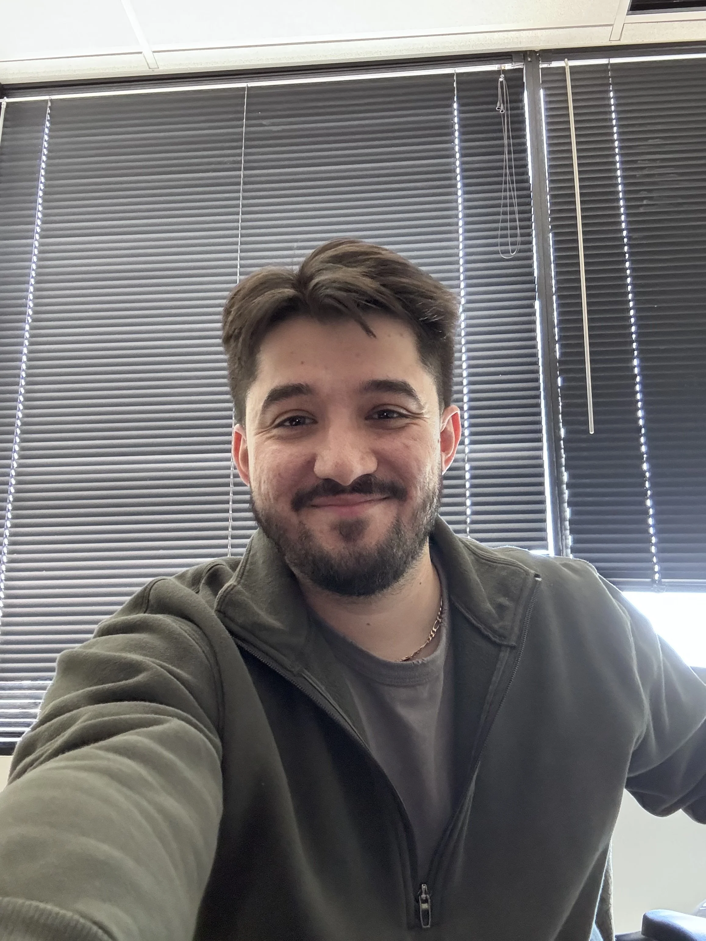 A smiling young man with brown hair and a beard taking a selfie indoors, with closed window blinds in the background.