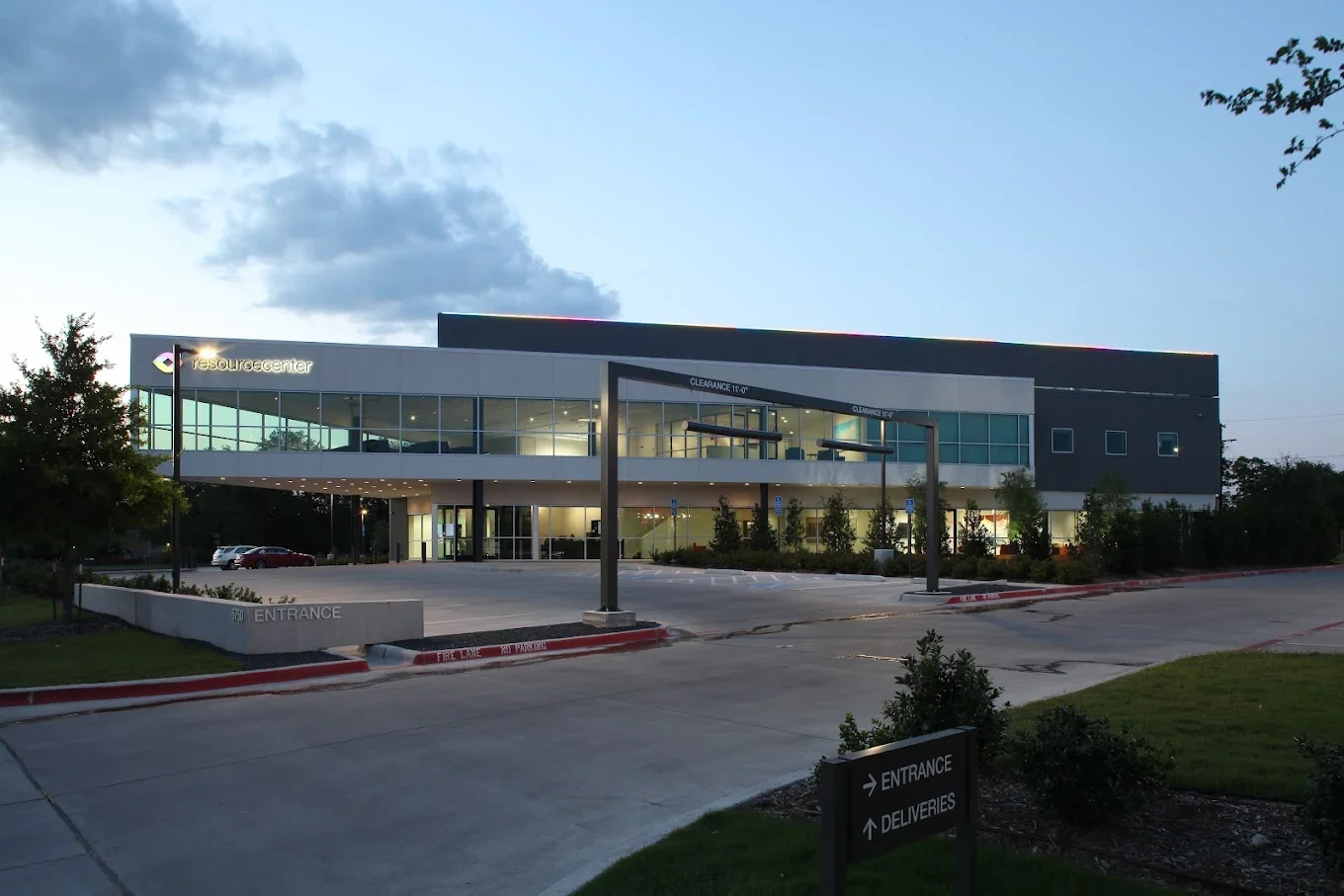 Modern two-story office building with glass windows and signage for Resource Center, parking lot in front, trees and landscaping around, early evening with cloudy sky.