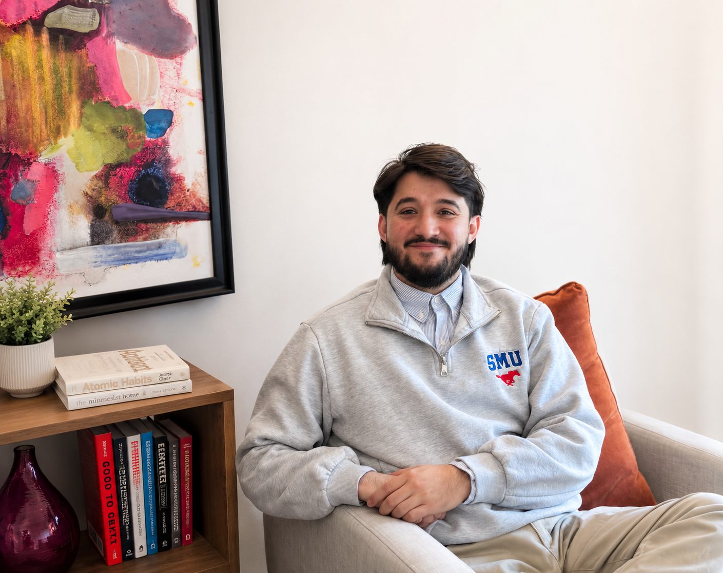 A young man with dark hair and a beard sitting on a beige armchair, wearing a gray sweatshirt with SMU logo, in a room with a white wall, a colorful painting, a wooden bookshelf with books and a plant, and a rust-colored cushion.