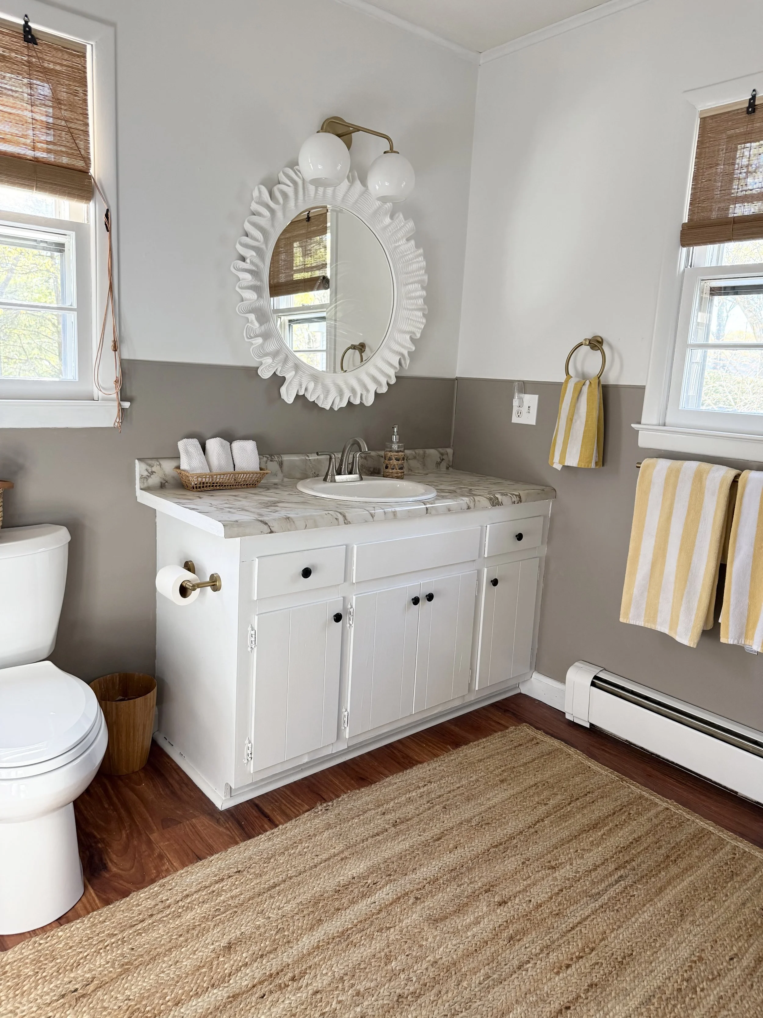 A bathroom with a white vanity, marble countertop, oval mirror with wavy white frame, two windows with bamboo blinds, a toilet, a brown wastebasket, and a beige area rug. There are yellow and white striped towels hanging on a brass towel ring and a towel bar.