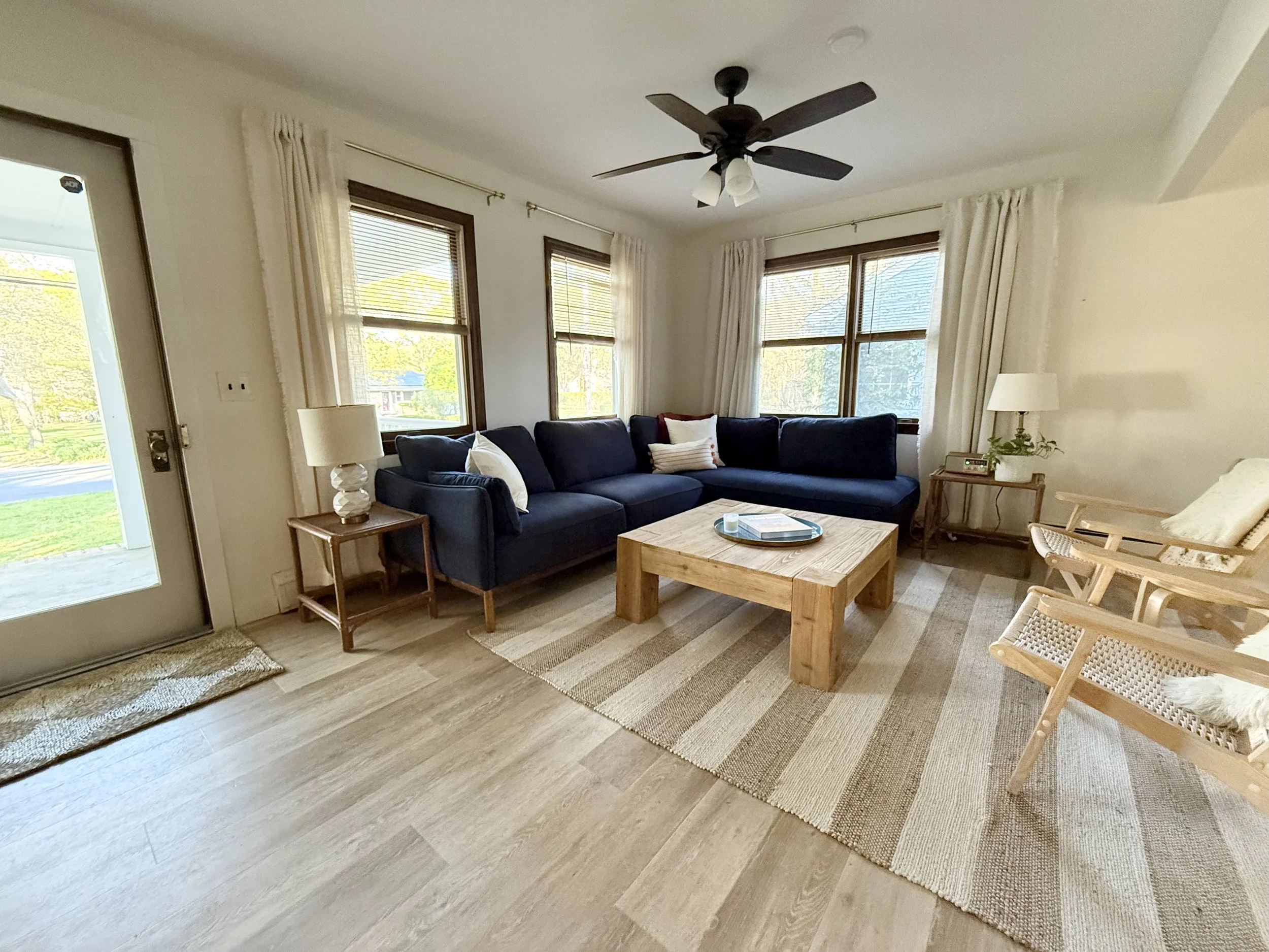 Living room with large windows, dark blue sectional sofa, wooden coffee table, side table with lamp, and wooden chairs with woven seats and cushions.