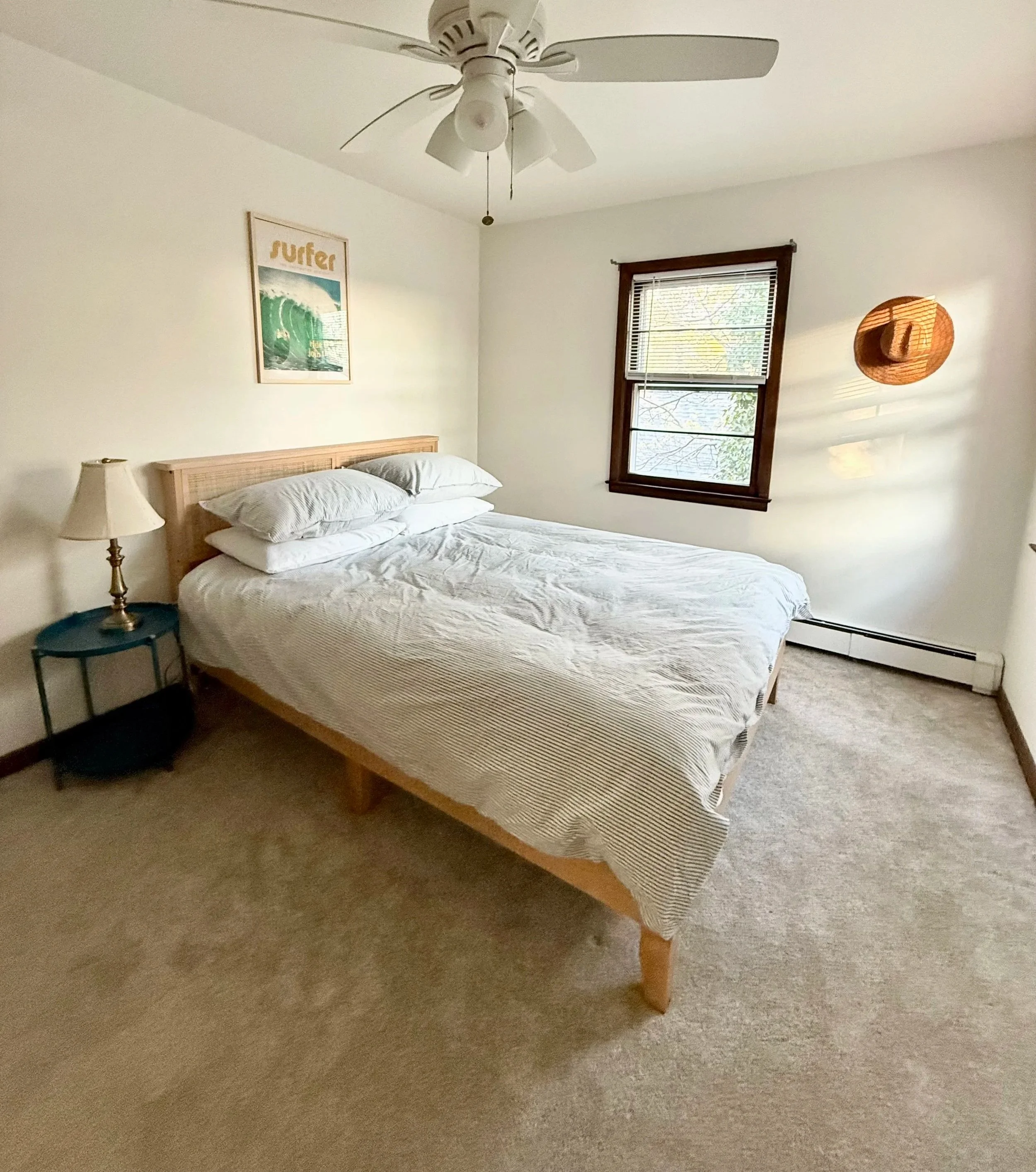 A bedroom with a wood-framed bed, white bedding, pillows, a blue side table with a brass lamp, a framed surf poster on the wall, a window with blinds, and a woven hat hanging on the wall.