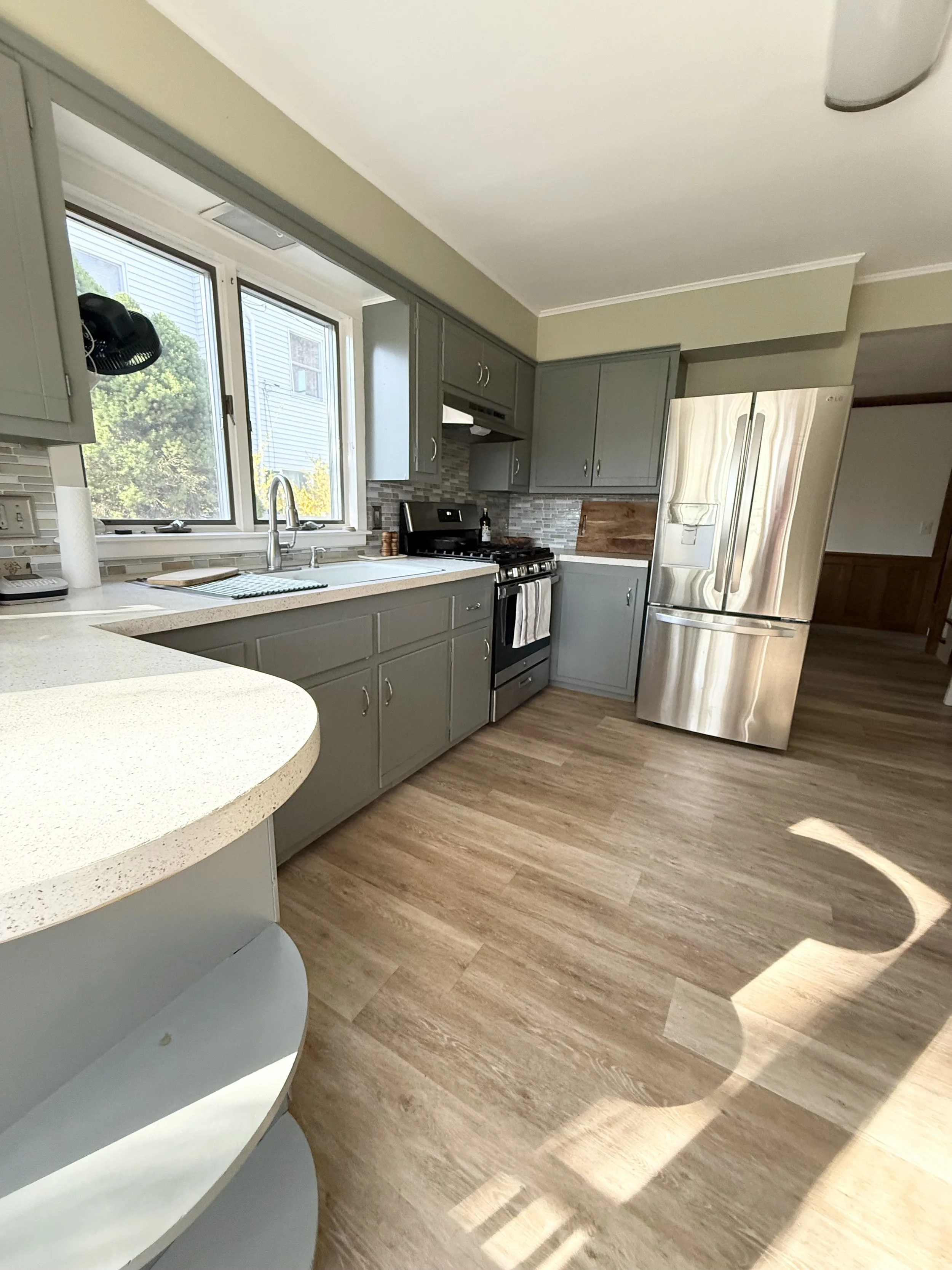 A kitchen with gray cabinets, a stainless steel refrigerator, a black stove, a beige countertop, a window with trees outside, and wooden flooring.