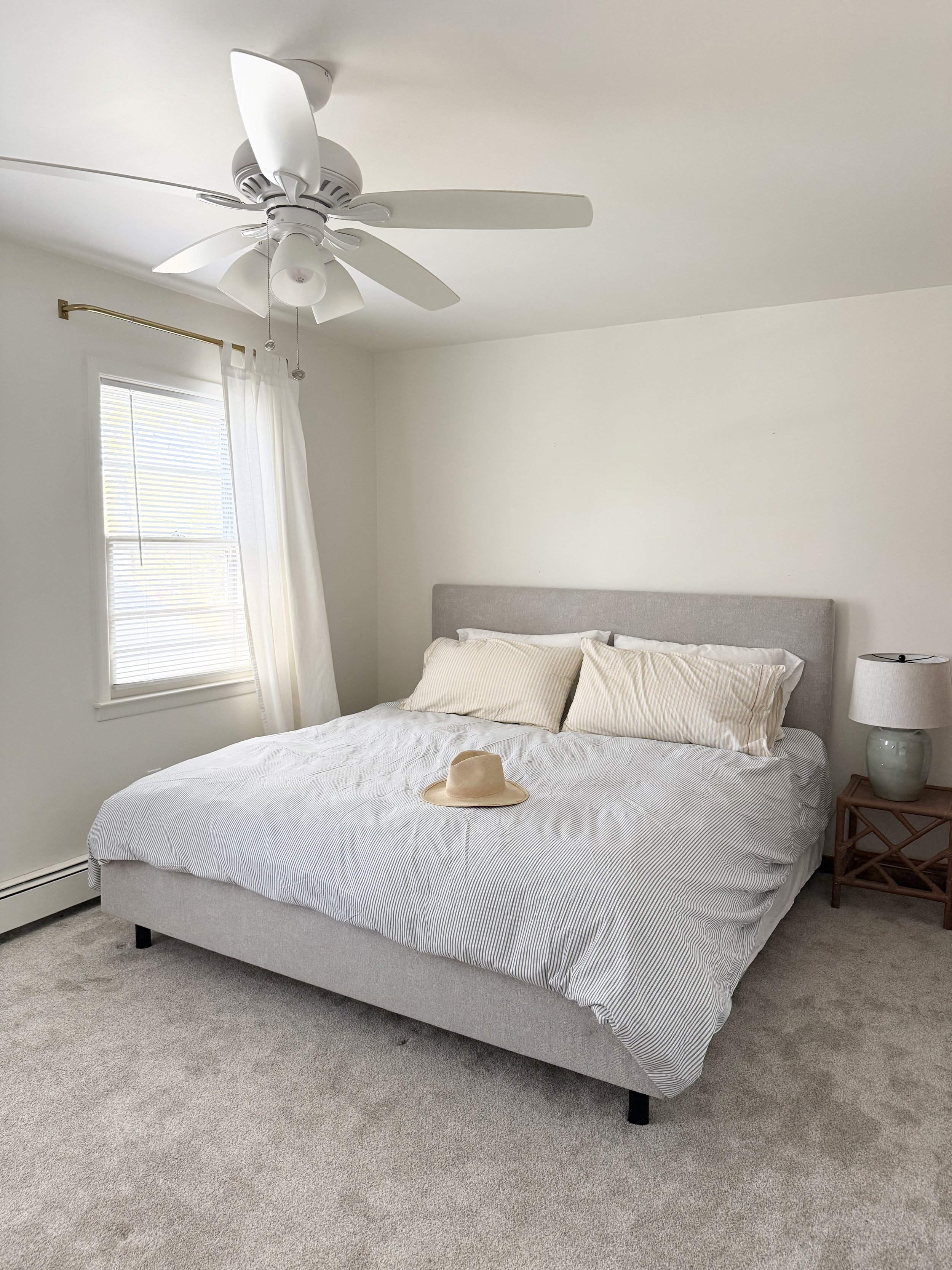 A bedroom with a bed, two pillows, a beige hat on the bed, a bedside table with a lamp, a window with white curtains, and a ceiling fan.