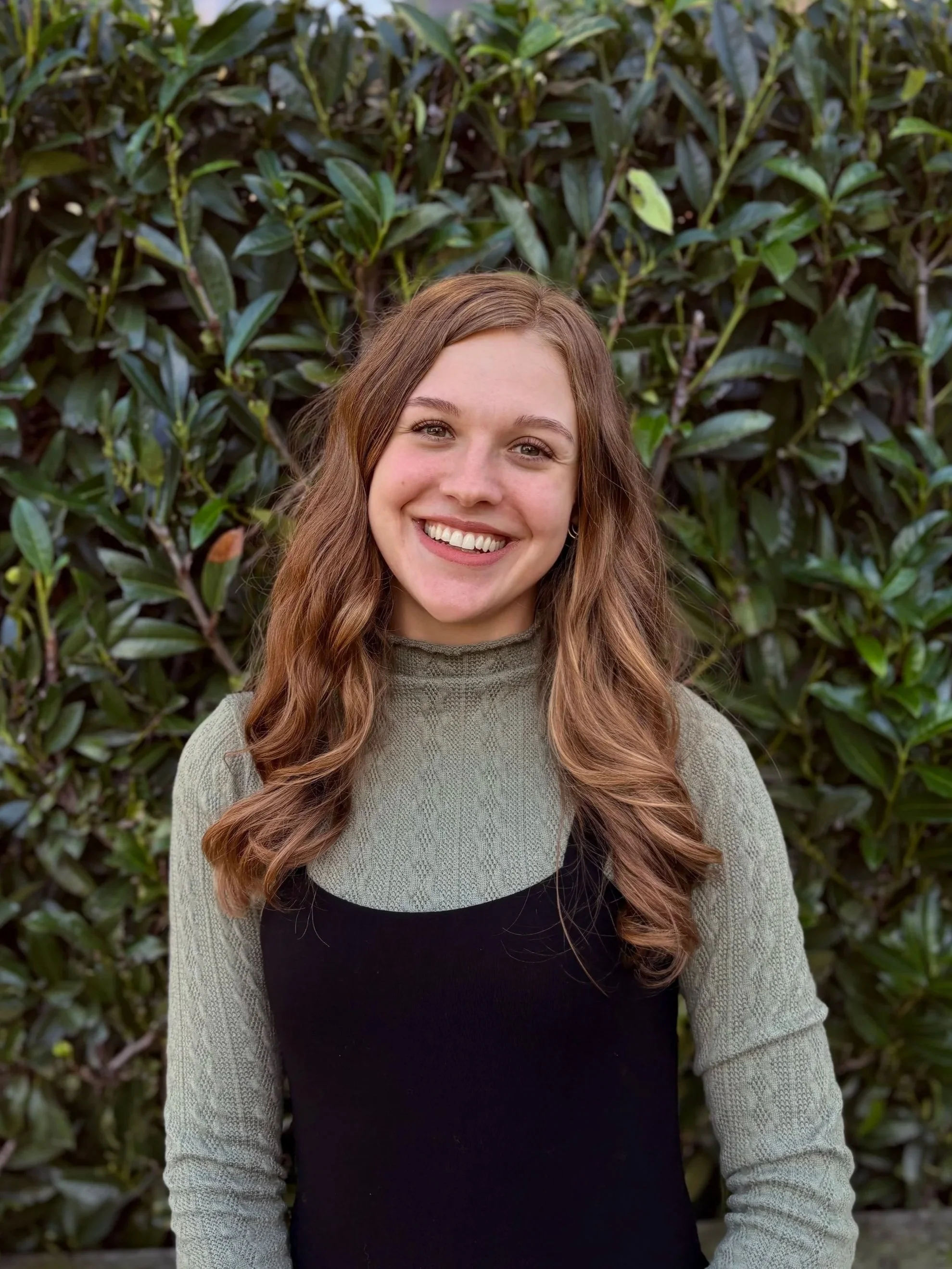 A young woman with long wavy brown hair smiling in front of green leafy bushes.