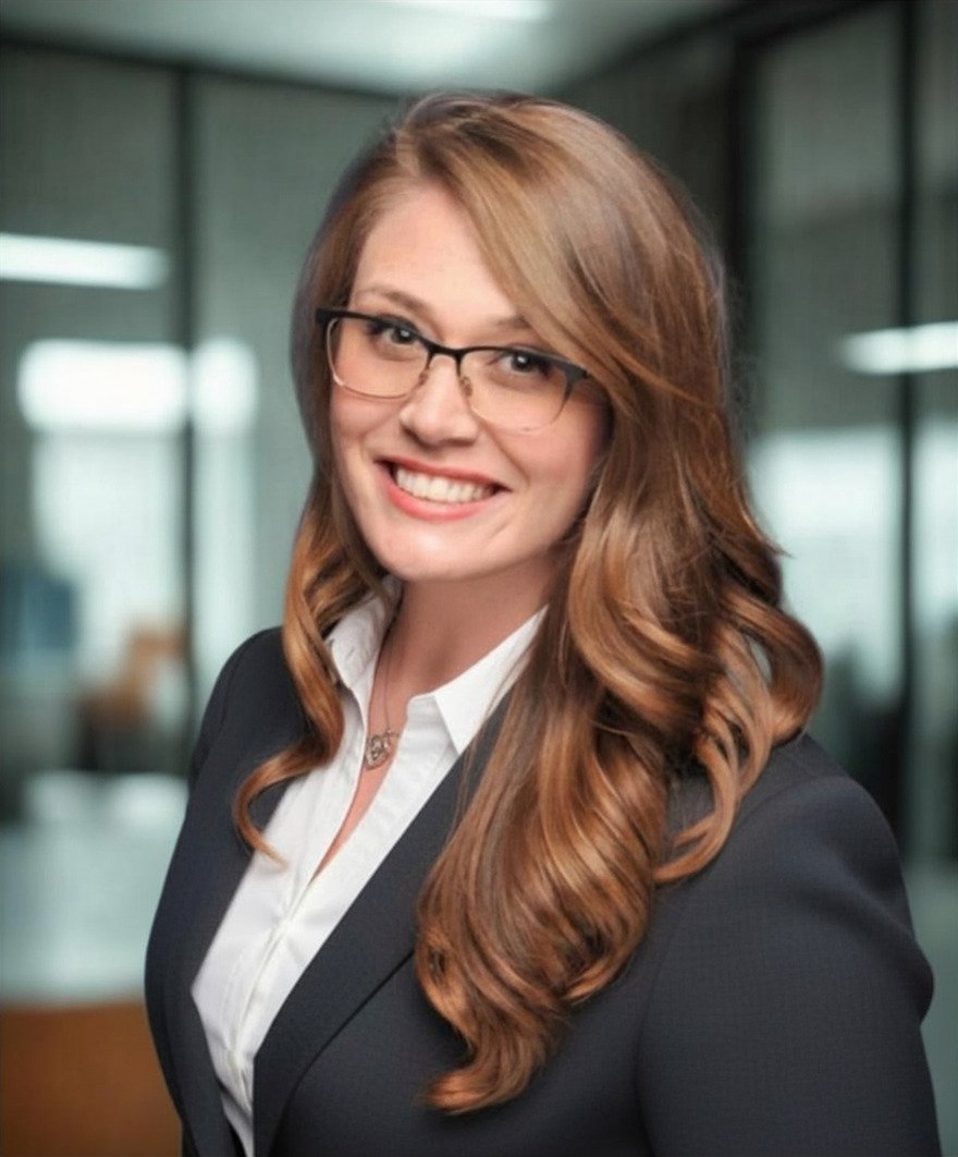 A woman with long, wavy red hair and glasses smiling in a professional setting, wearing a dark blazer and white collared shirt.