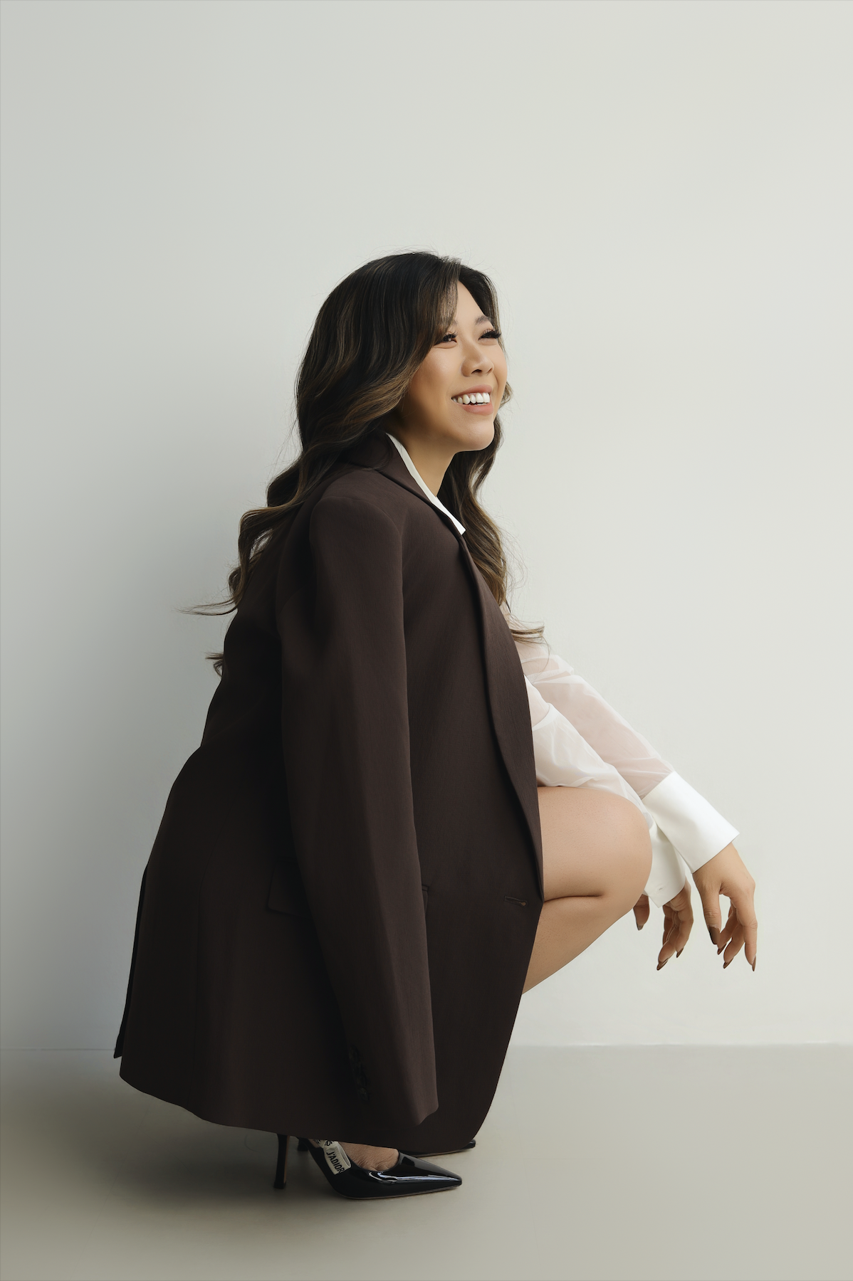 A woman with long wavy hair wearing a brown blazer and white shirt, smiling and squatting with her right knee raised, in front of a plain light-colored wall.