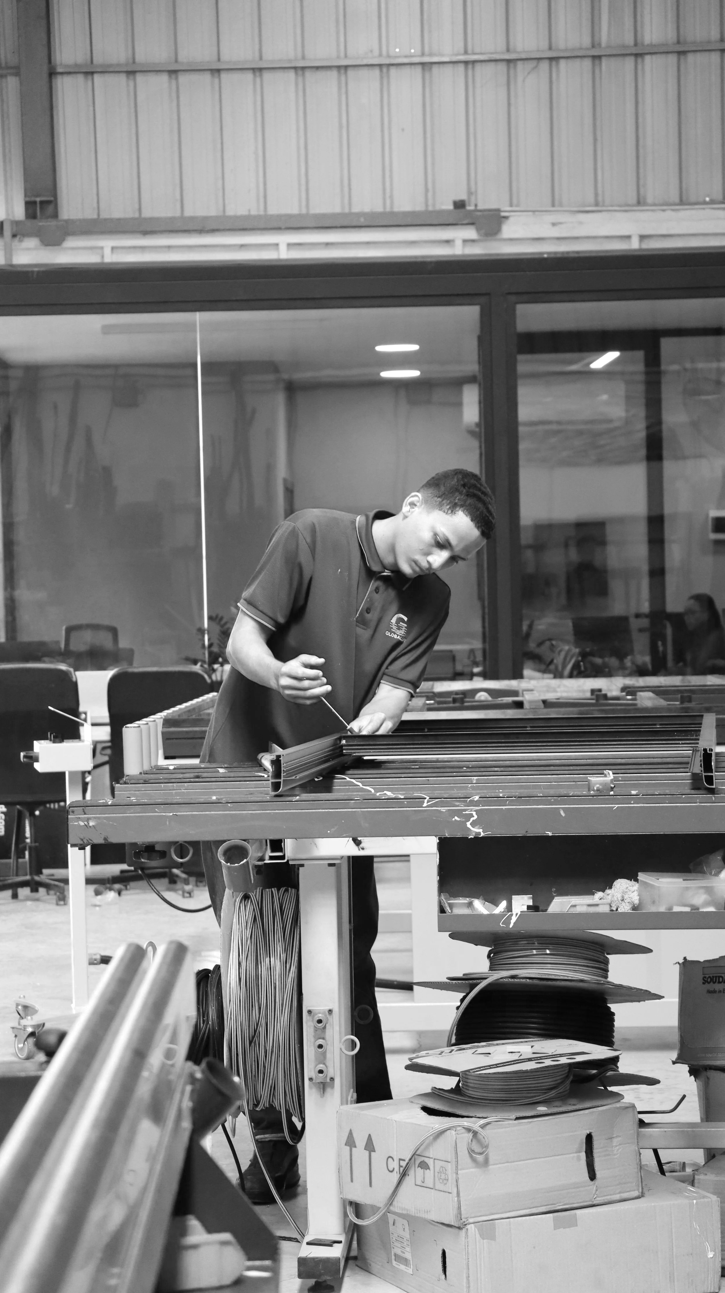 A man working in an industrial or workshop setting, examining or assembling a piece of equipment at a workbench surrounded by various tools and supplies.