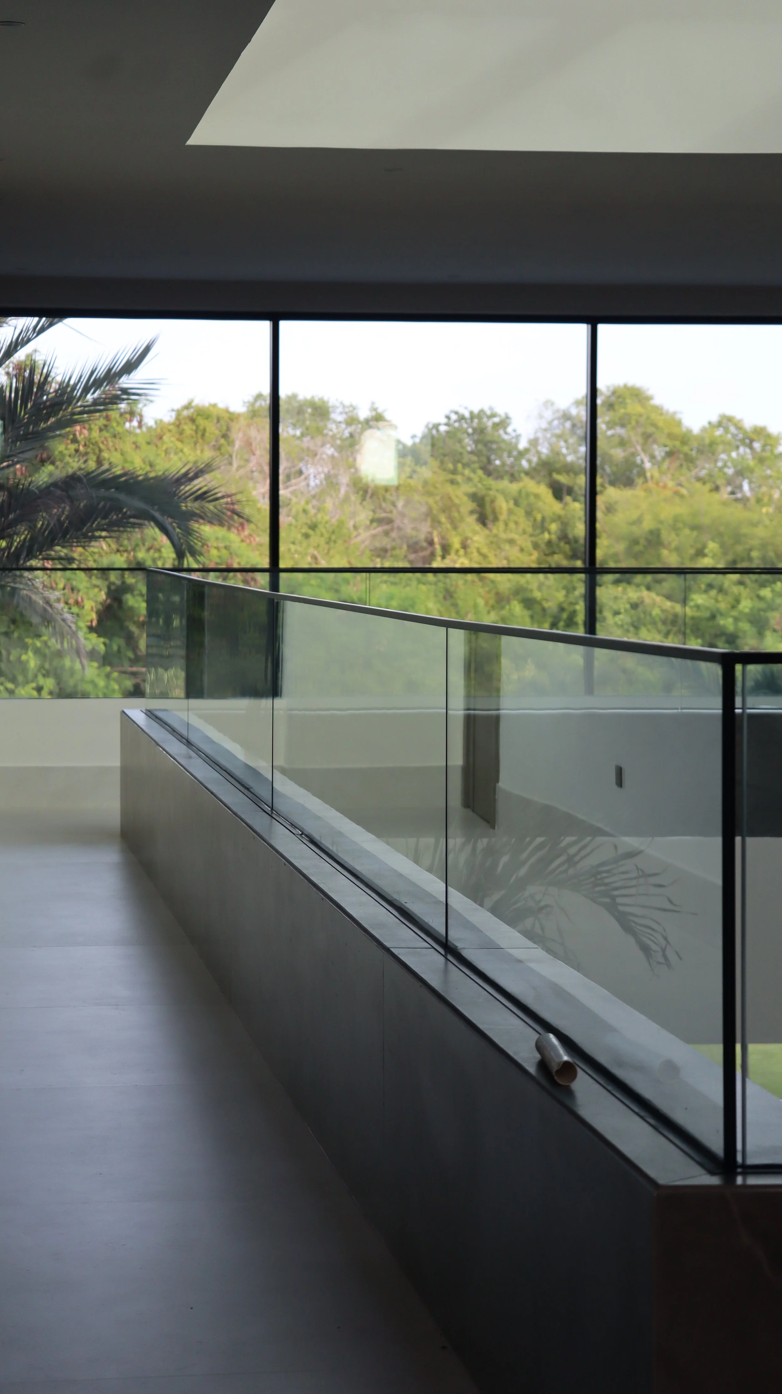 Interior view of a modern building with large floor-to-ceiling windows showing green trees outside, a glass railing along a staircase or balcony, and a plant partially visible on the left side.