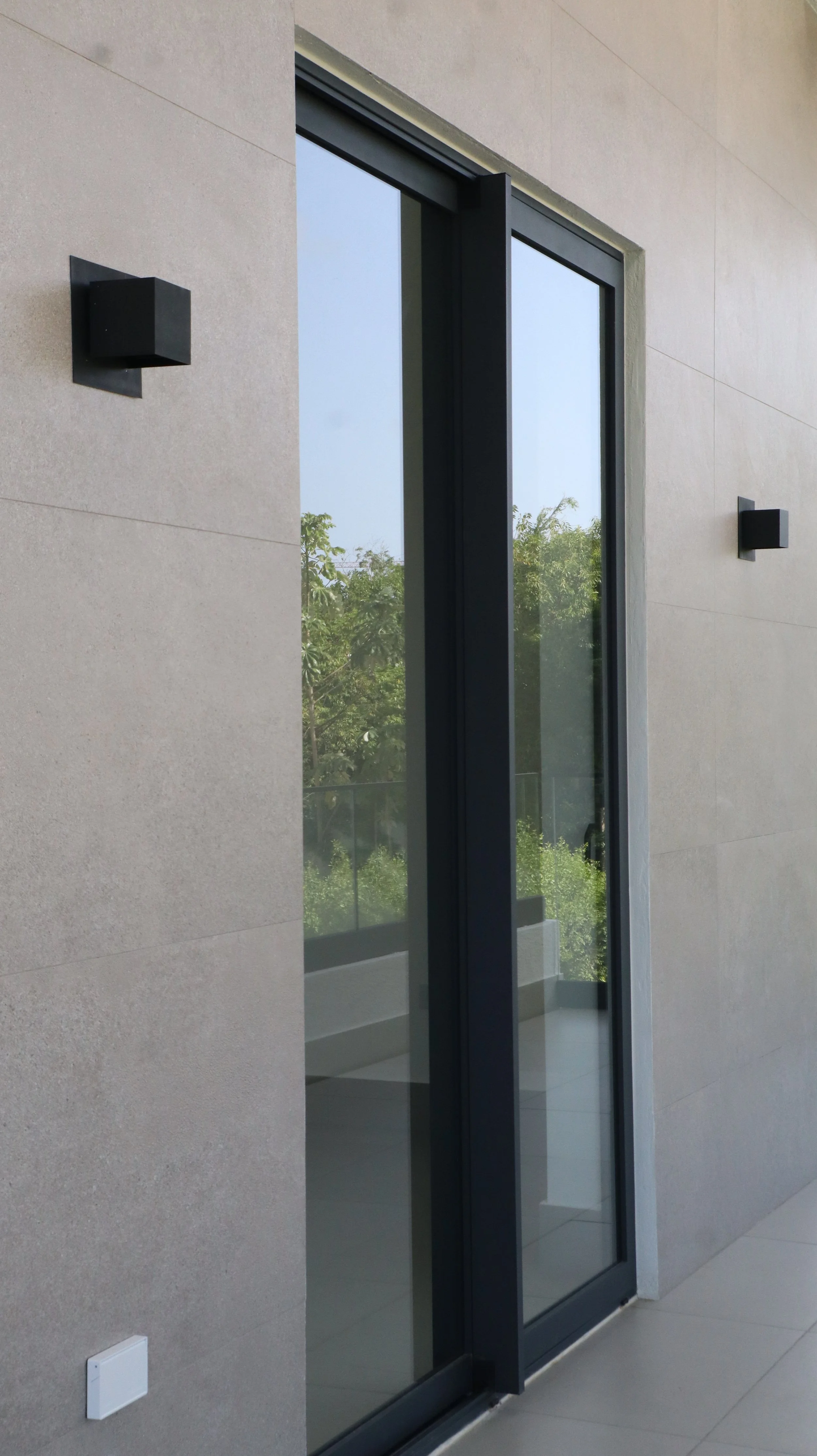 Modern sliding glass door with black frame and beige tiled wall, featuring two black outdoor wall lamps, and a view of greenery outside.