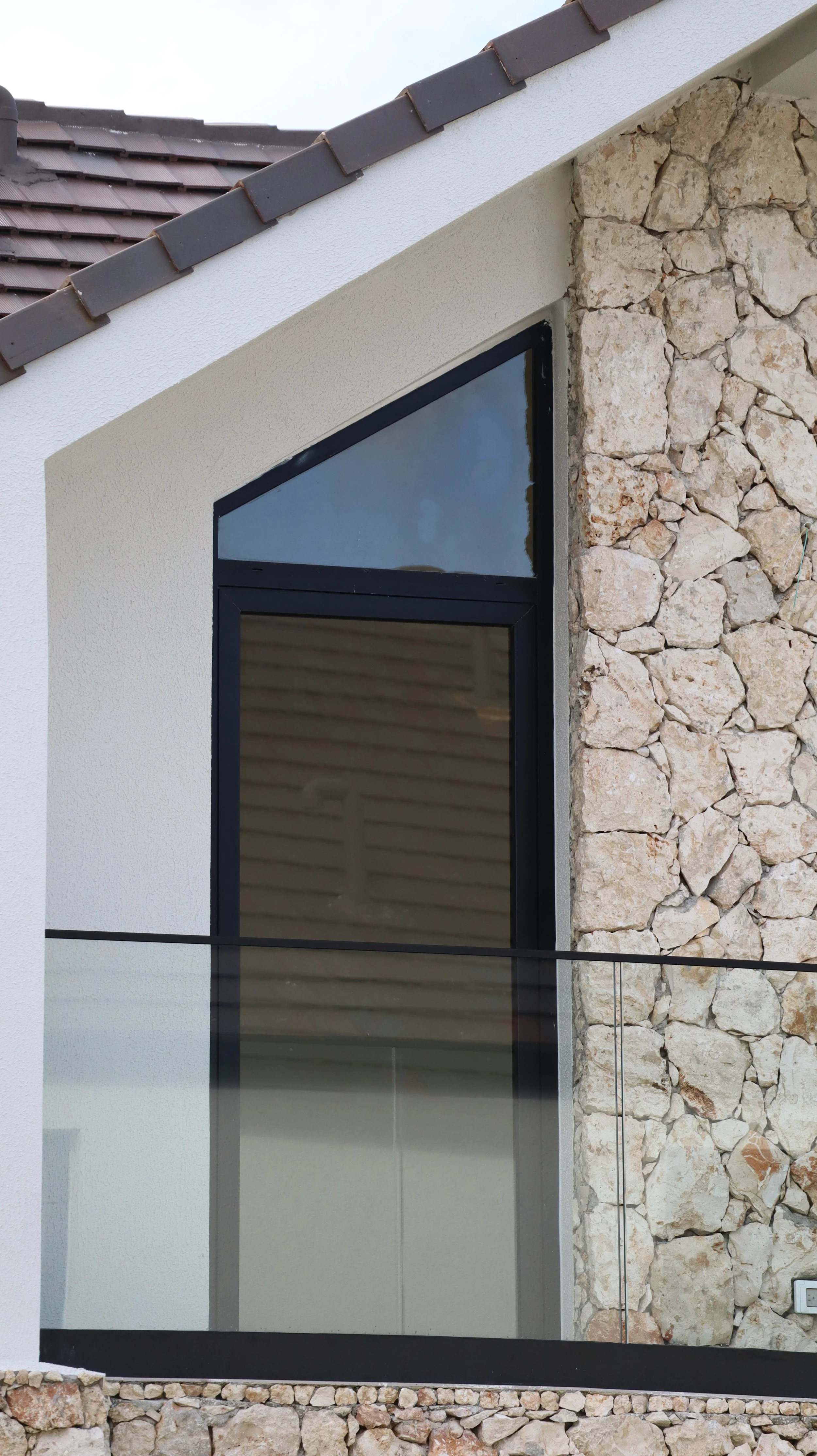 Close-up of a modern house exterior with a large glass window, white stucco wall on the left, and a stone wall on the right. The roof has brown tiles.