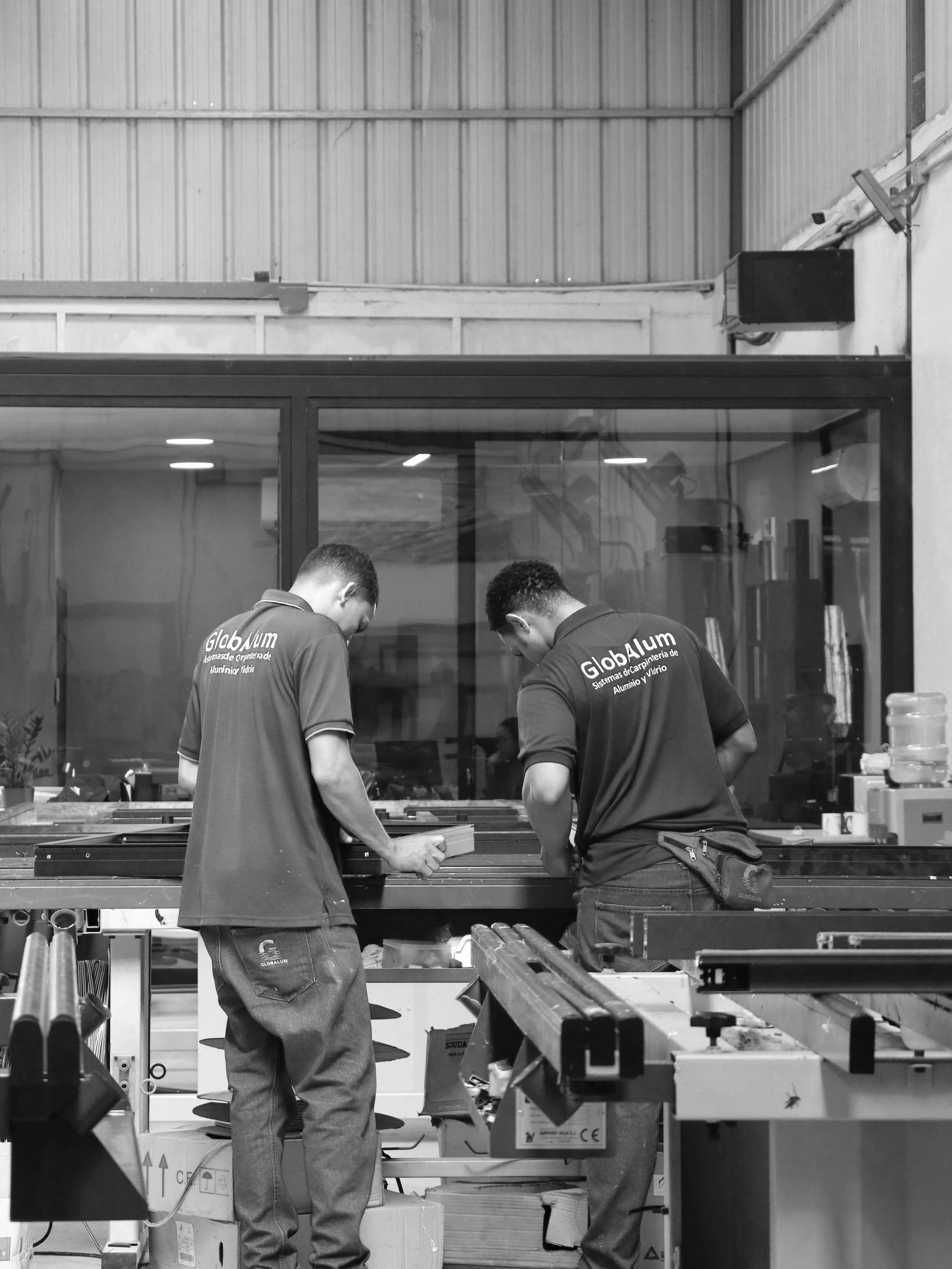 Two workers wearing shirts with 'Globalum' logo working at a table in a workshop or factory setting, surrounded by tools and materials.