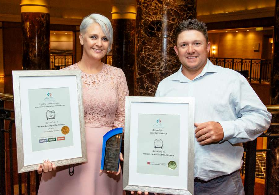 A woman and a man standing indoors, holding framed awards. The woman has short white hair and is wearing a pink lace dress. The man has short brown hair and is wearing a light blue dress shirt. They are smiling for the photo in a warmly lit area with marble pillars and a staircase in the background.