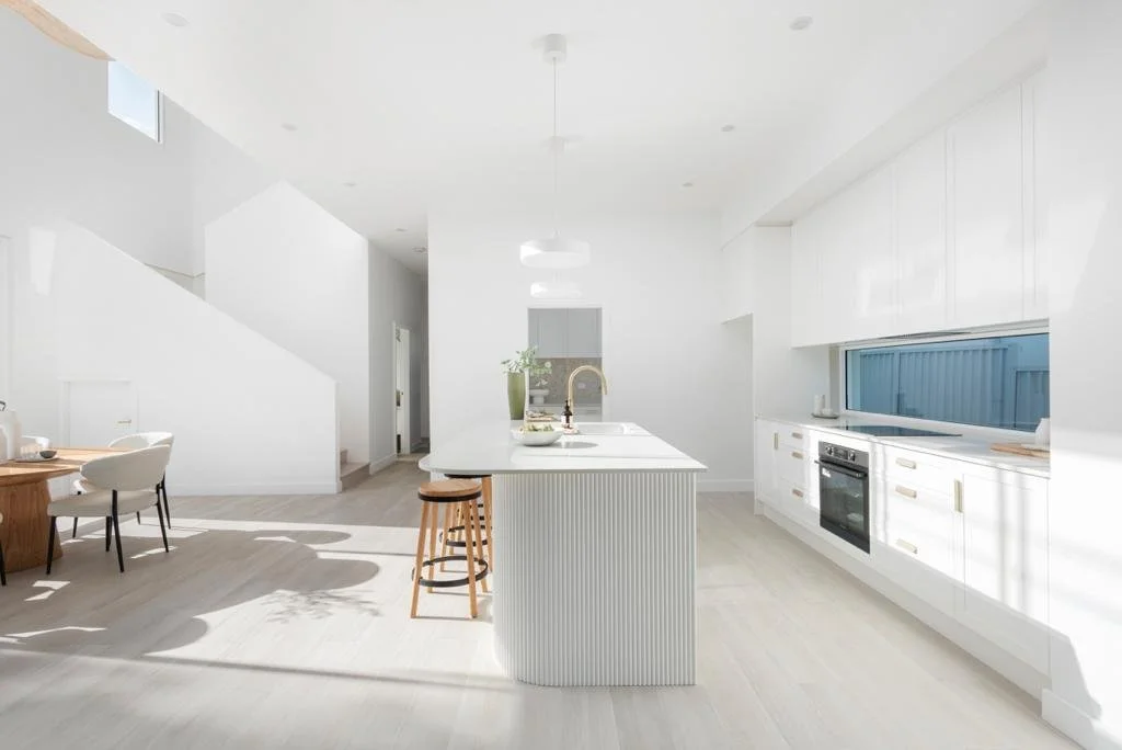 Bright, white kitchen with a central island, built-in oven, and large window.