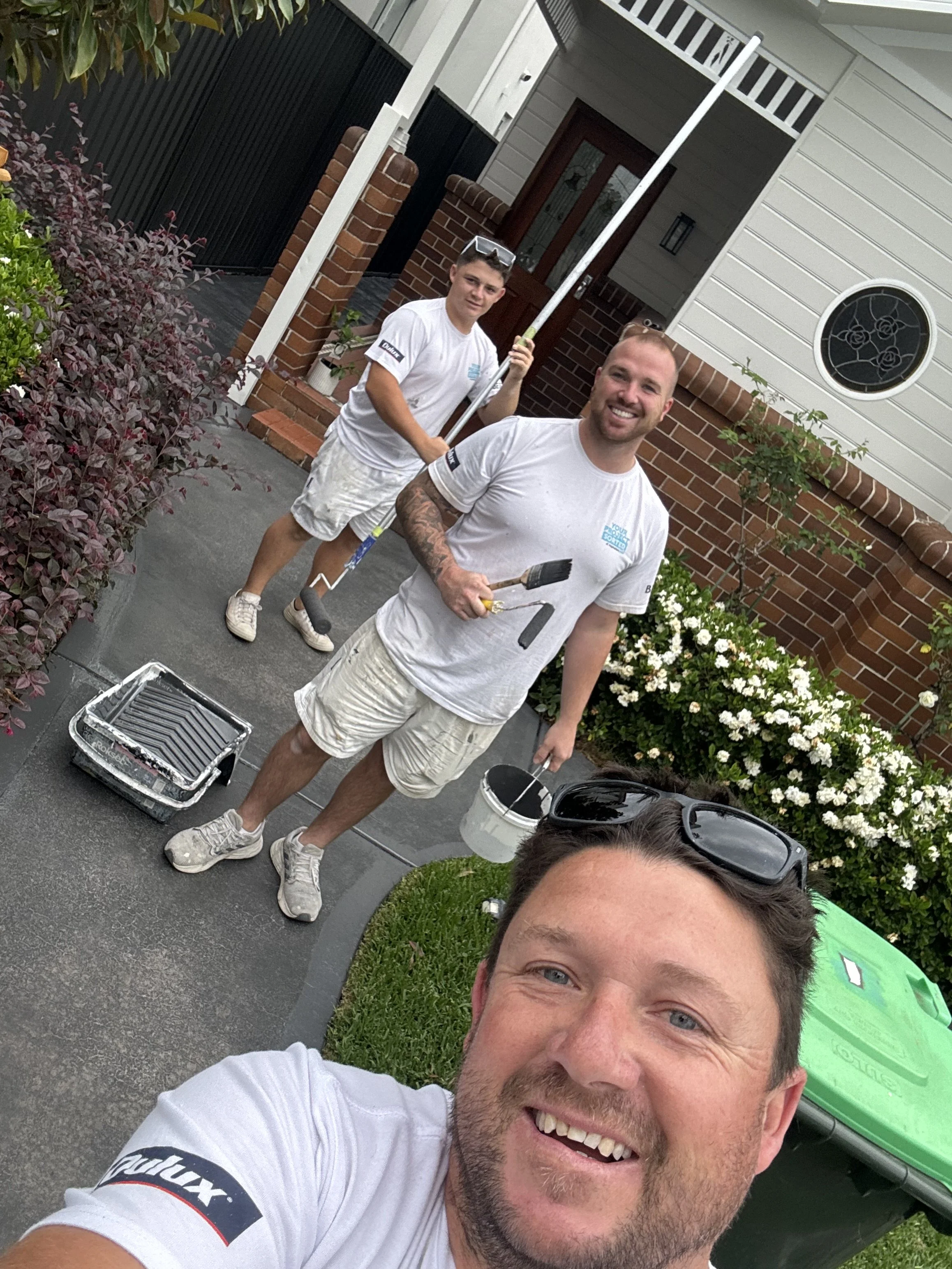 Three men are smiling on a freshly painted porch in front of a house. The man in the foreground is taking a selfie, with three others behind him holding painting tools and a bucket of paint.