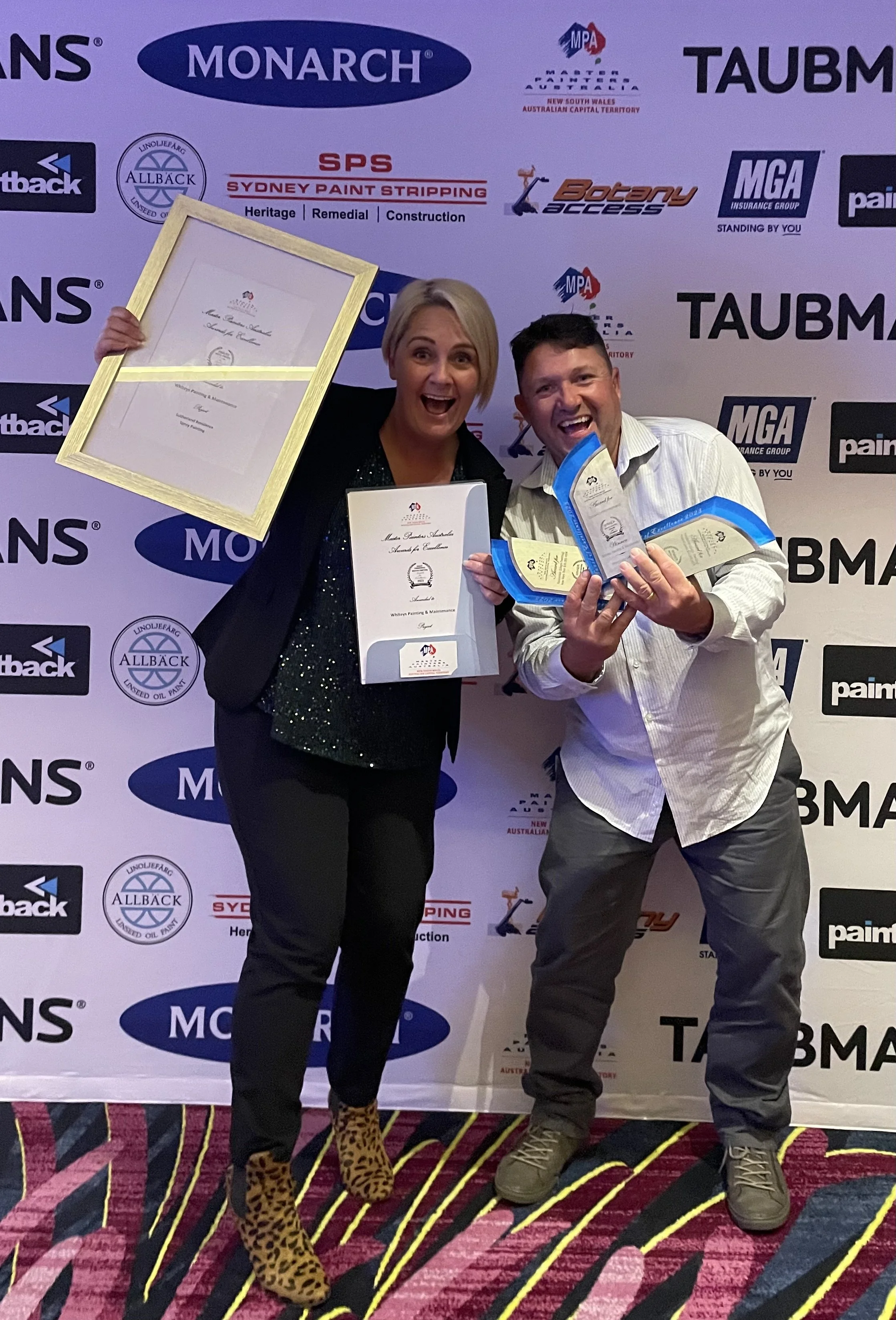 Two people celebrating and holding awards in front of a backdrop with various company logos at a corporate event.