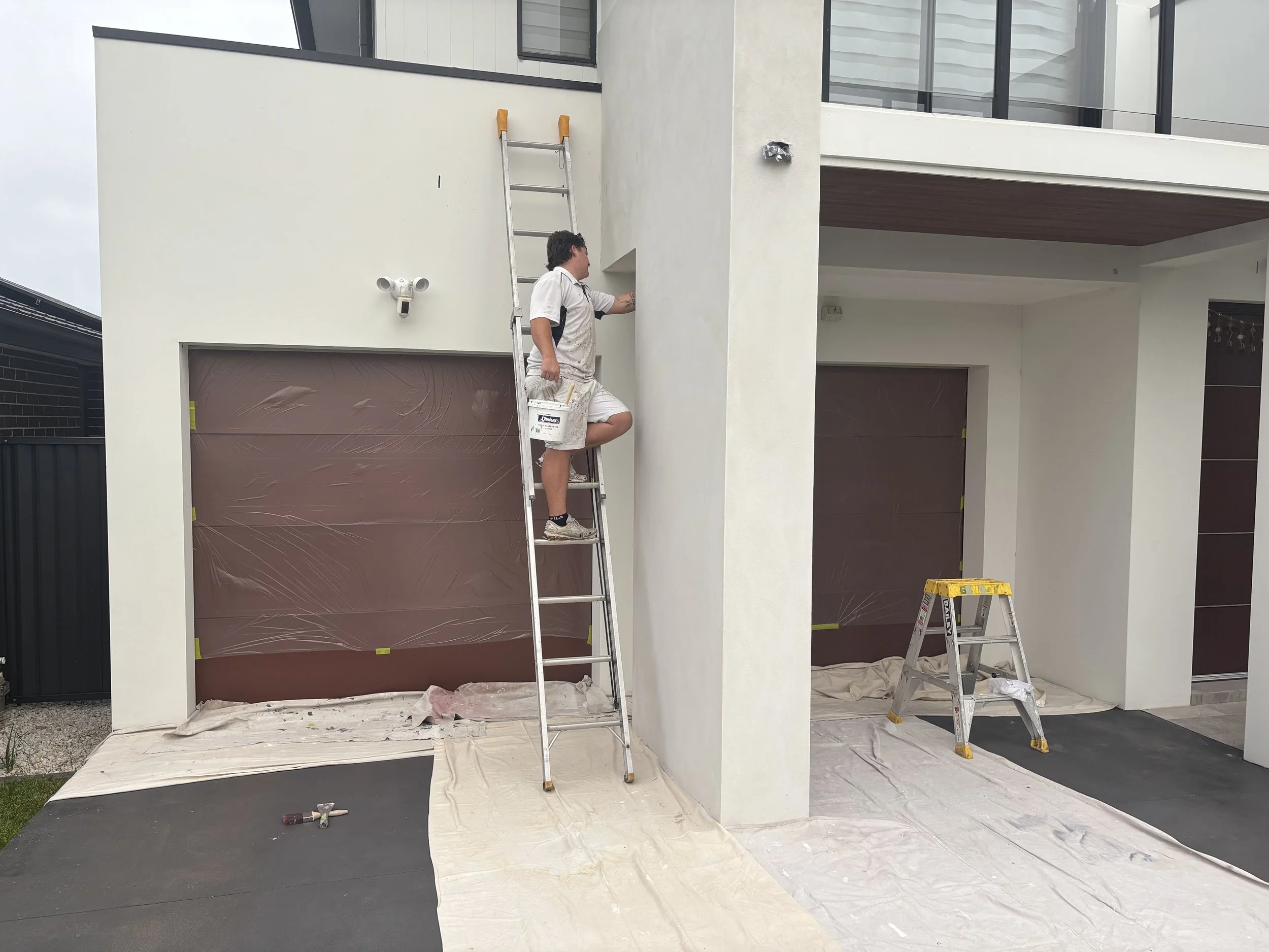 A person standing on a ladder painting the exterior wall of a modern house, with painter's tools and protective coverings visible.