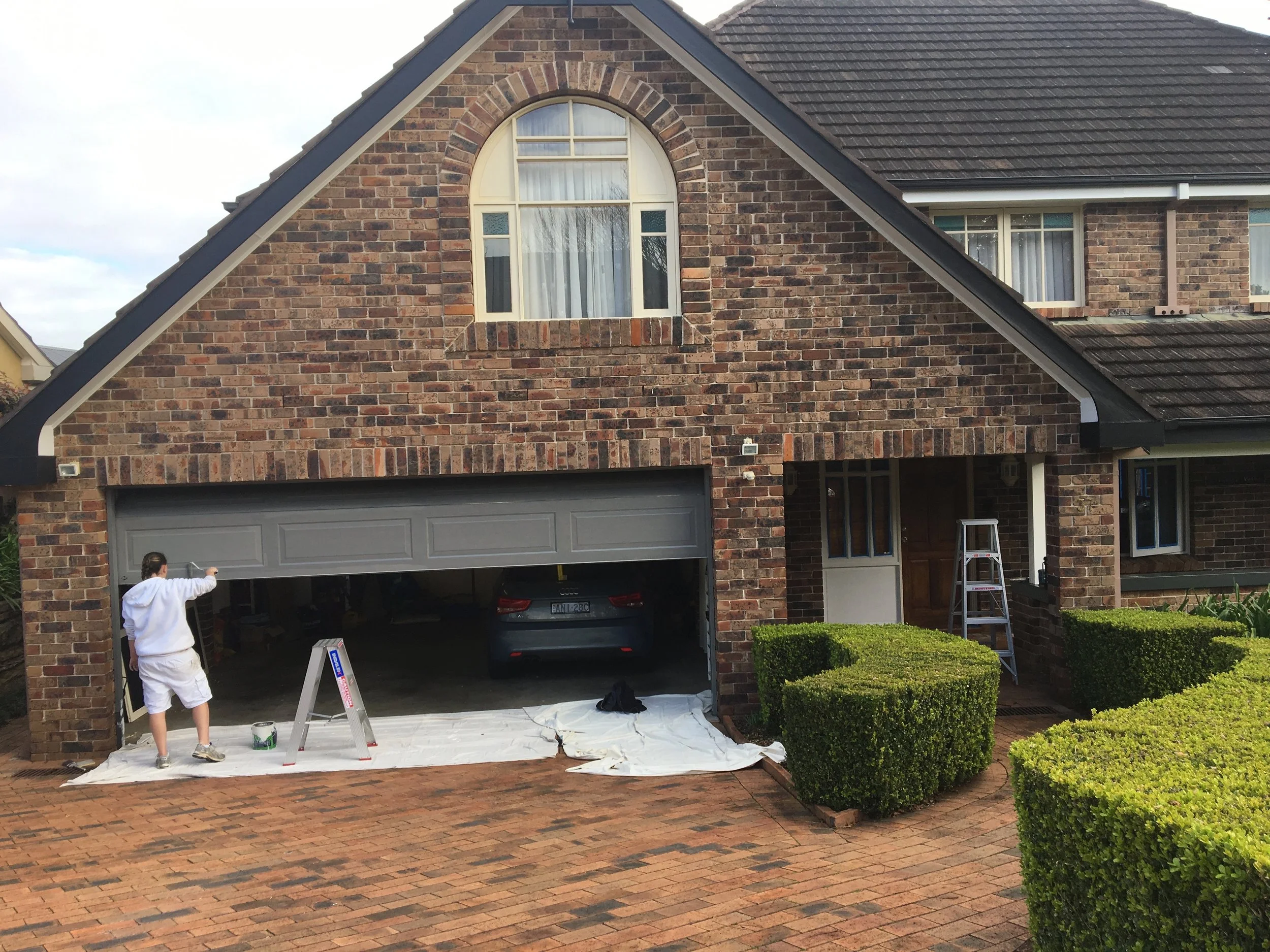 Painter in Sydney, NSW, painting the garage door of a brick house, with a ladder and painting supplies nearby, and a black car parked inside the garage.