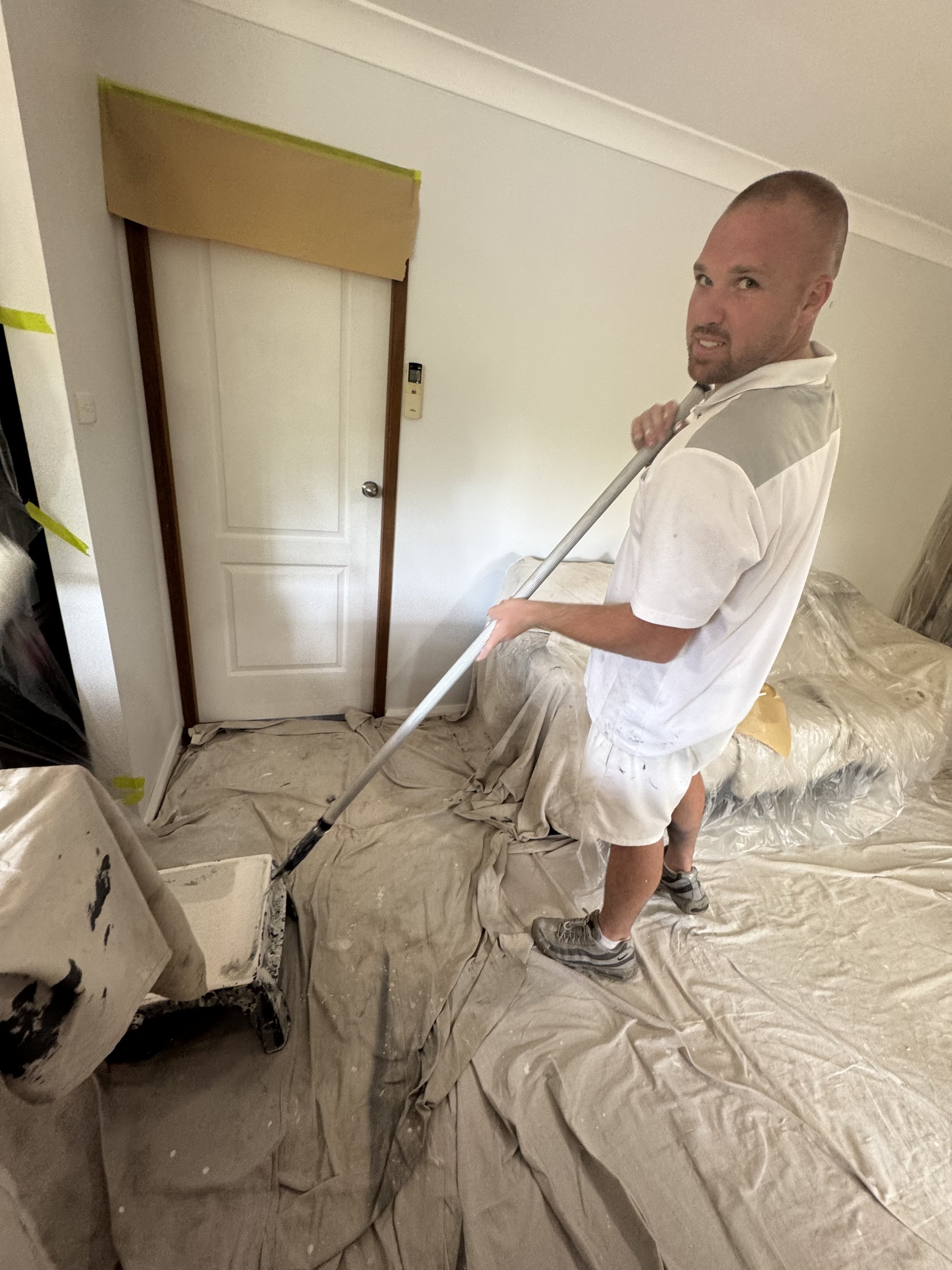 A man standing on a covered floor, using a paint roller with a long handle to paint the wall or ceiling. The room appears to be in the process of renovation, with plastic sheets covering the furniture and floor.