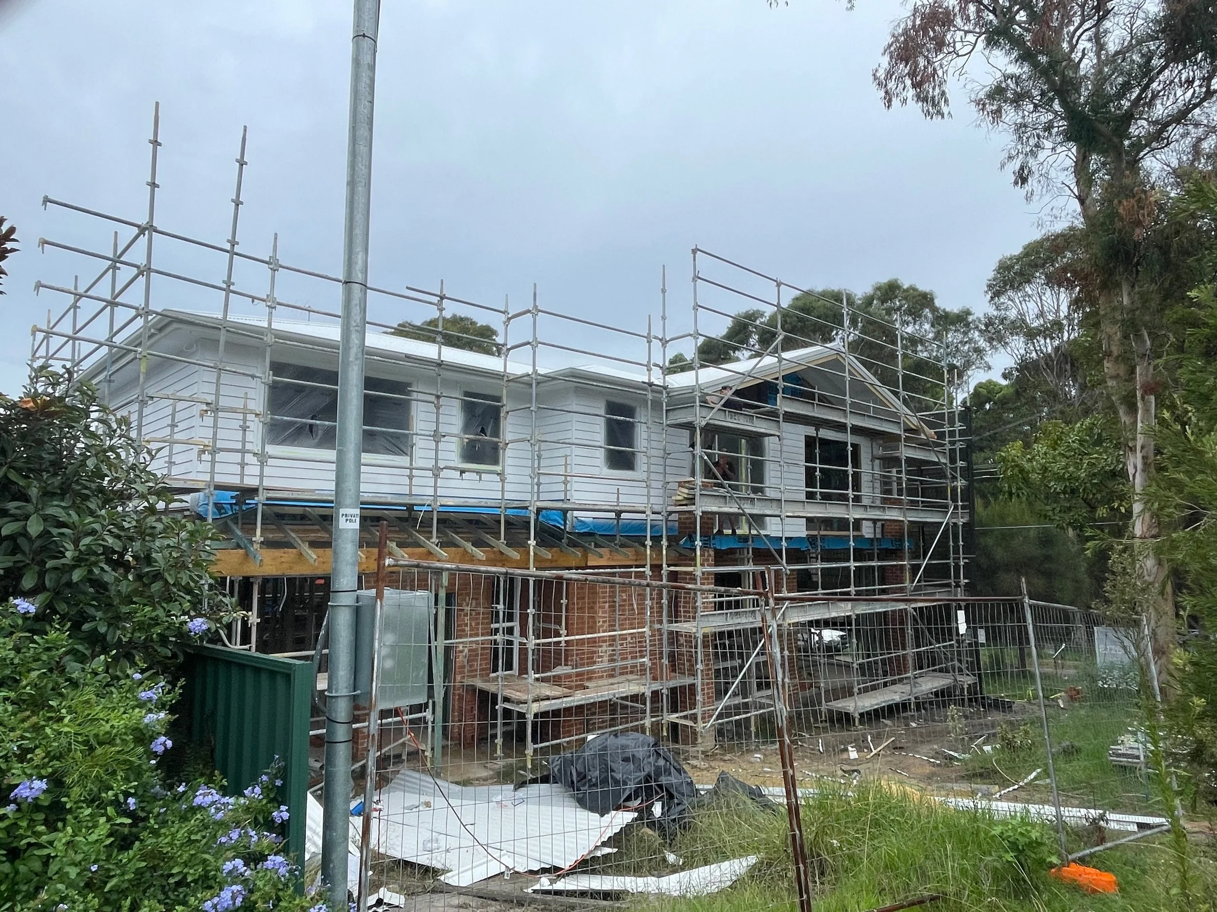 House under construction with scaffolding around it, trees and greenery in the background, and construction materials on the ground.