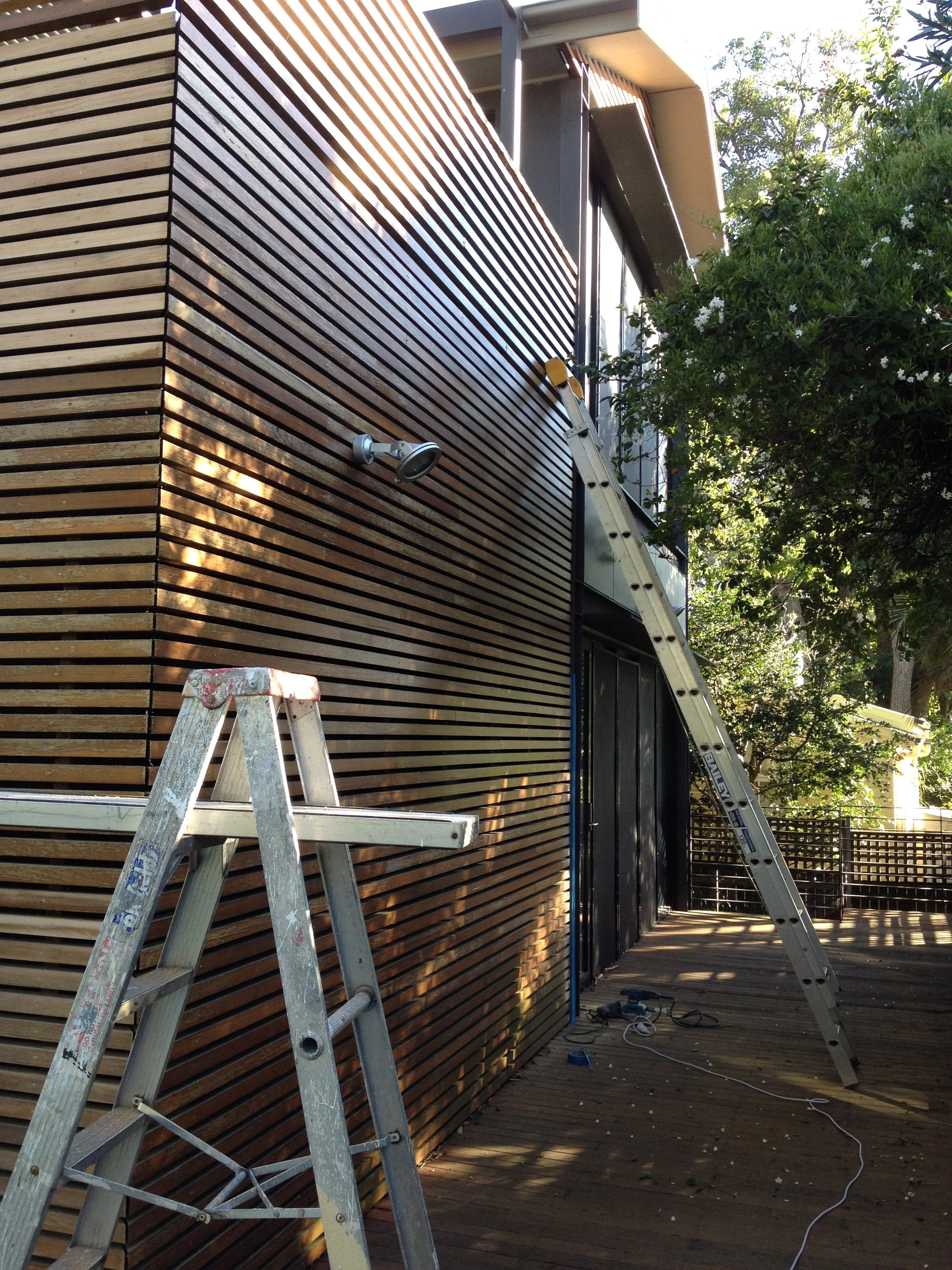 Construction or renovation work on a modern wooden building with a ladder leaning against the wall and a work light attached, surrounded by trees.