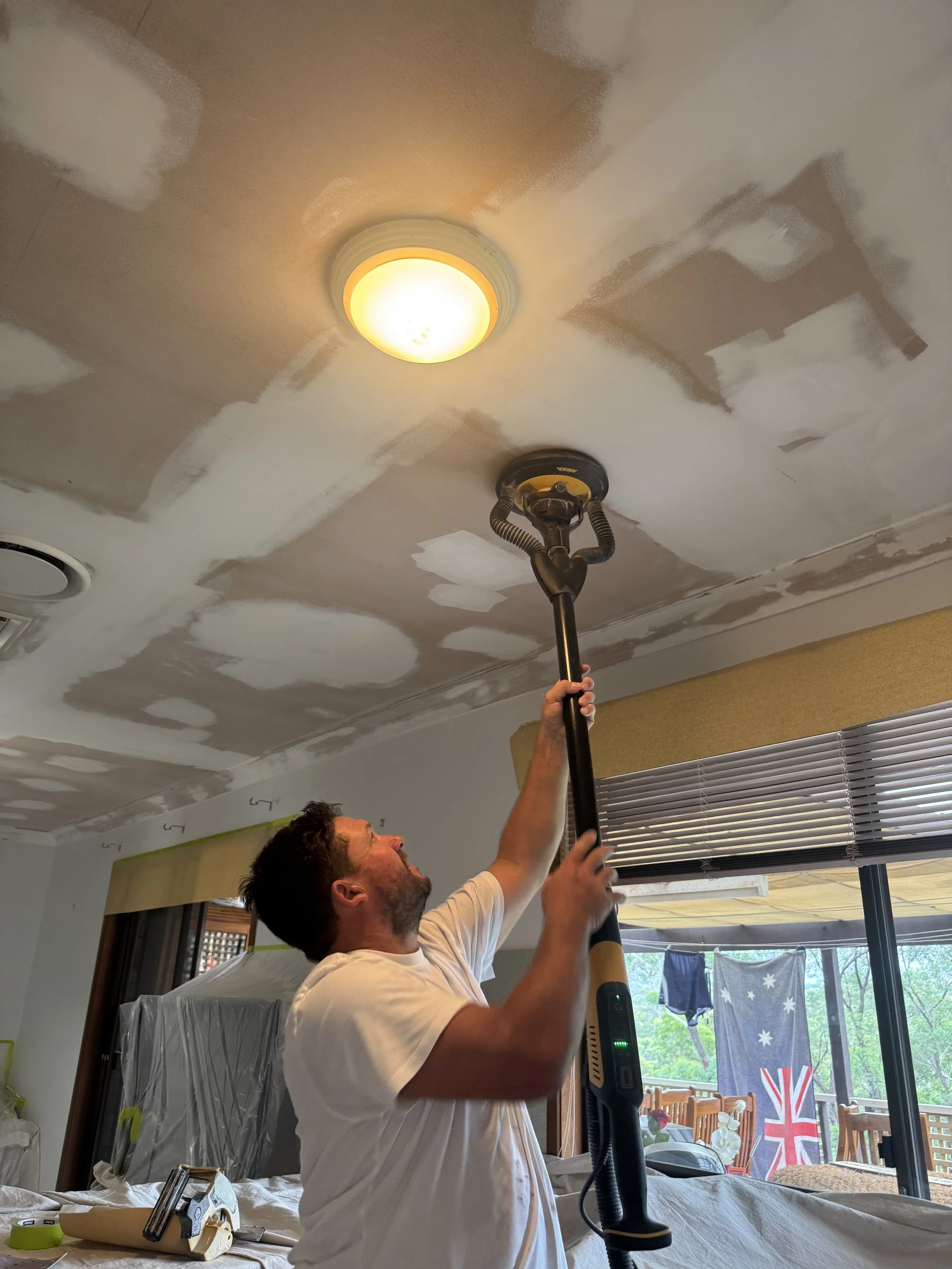 A man is using a drywall sander on a ceiling with patches of joint compound during home renovation.