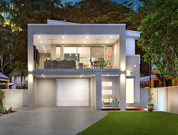 Modern two-story house with illuminated living room and garage, surrounded by trees and a lawn at dusk.