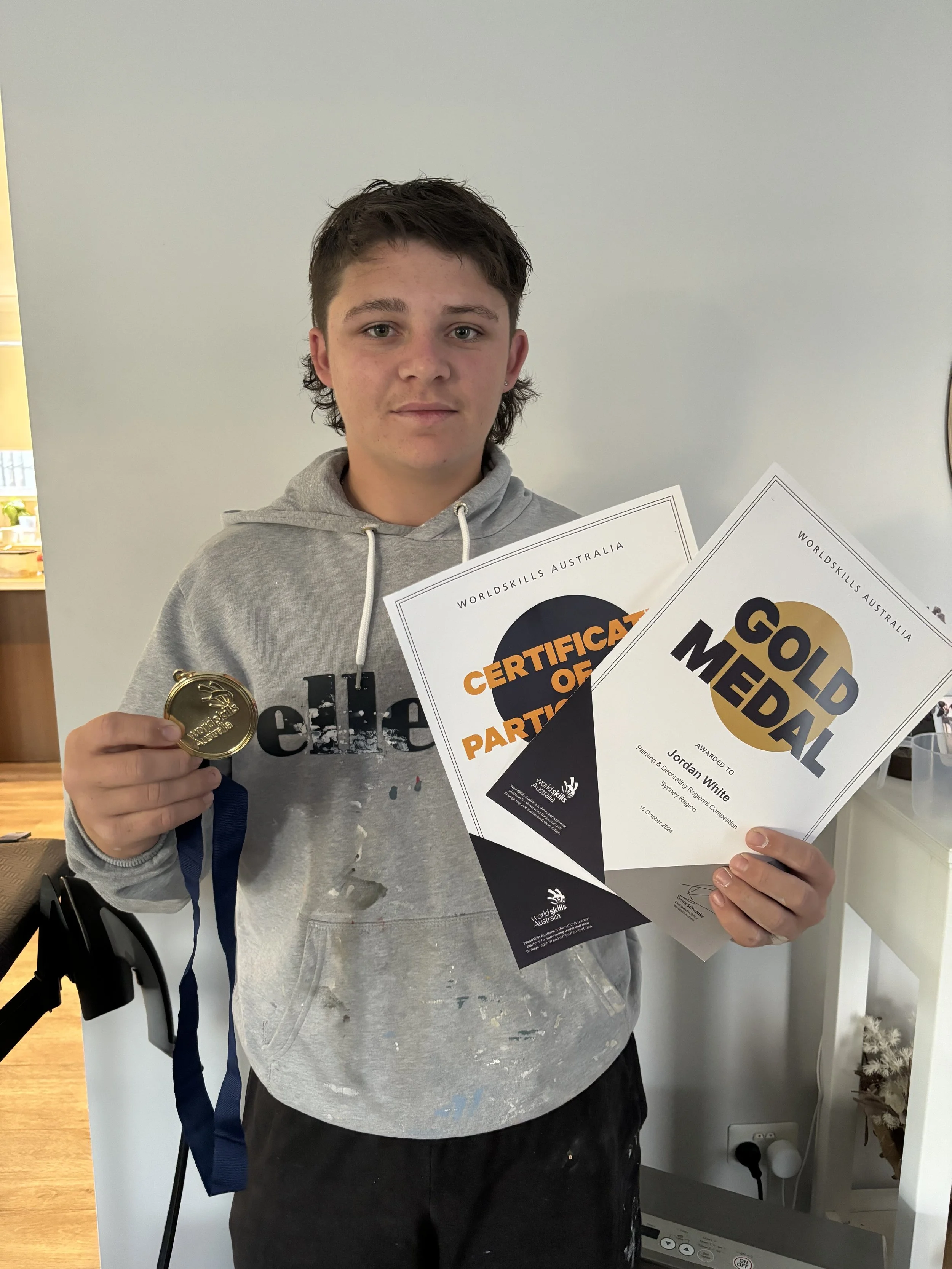 Young boy holding a gold medal, certificates for participation and a gold medal award in a room with a white wall.