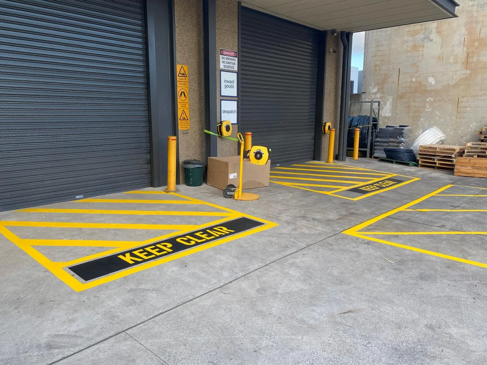 Clear yellow and black parking lot markings with the words "KEEP CLEAR" outlined on the ground in front of a warehouse loading dock with black roll-up doors and safety signs. Line Marking, painting for line marking services
