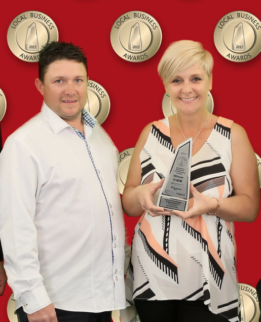 A woman holding a Local Business Award trophy standing next to a man against a red background with multiple awards plaques.