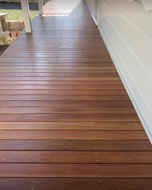 Wooden porch deck attached to a house, with a white exterior wall and a grassy yard visible in the background.