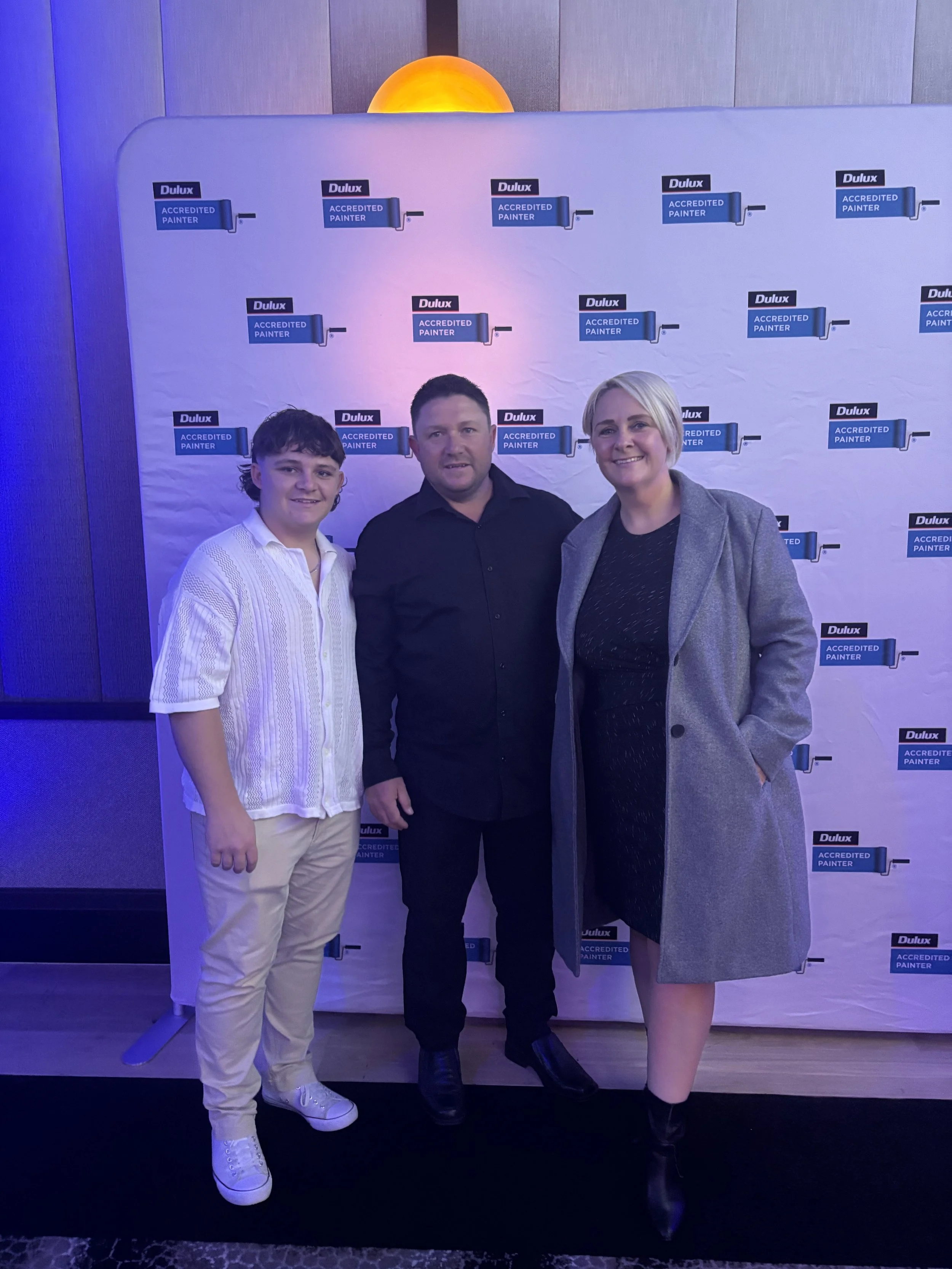 Three people standing in front of a backdrop with logos of Dulux and 'Accredited Painter'.