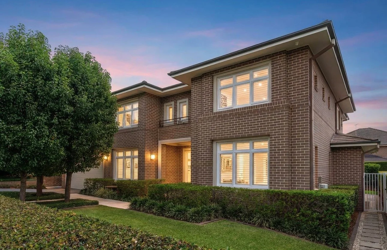 A two-story brick house with lit windows at dusk, surrounded by a well-maintained lawn, bushes, and trees.