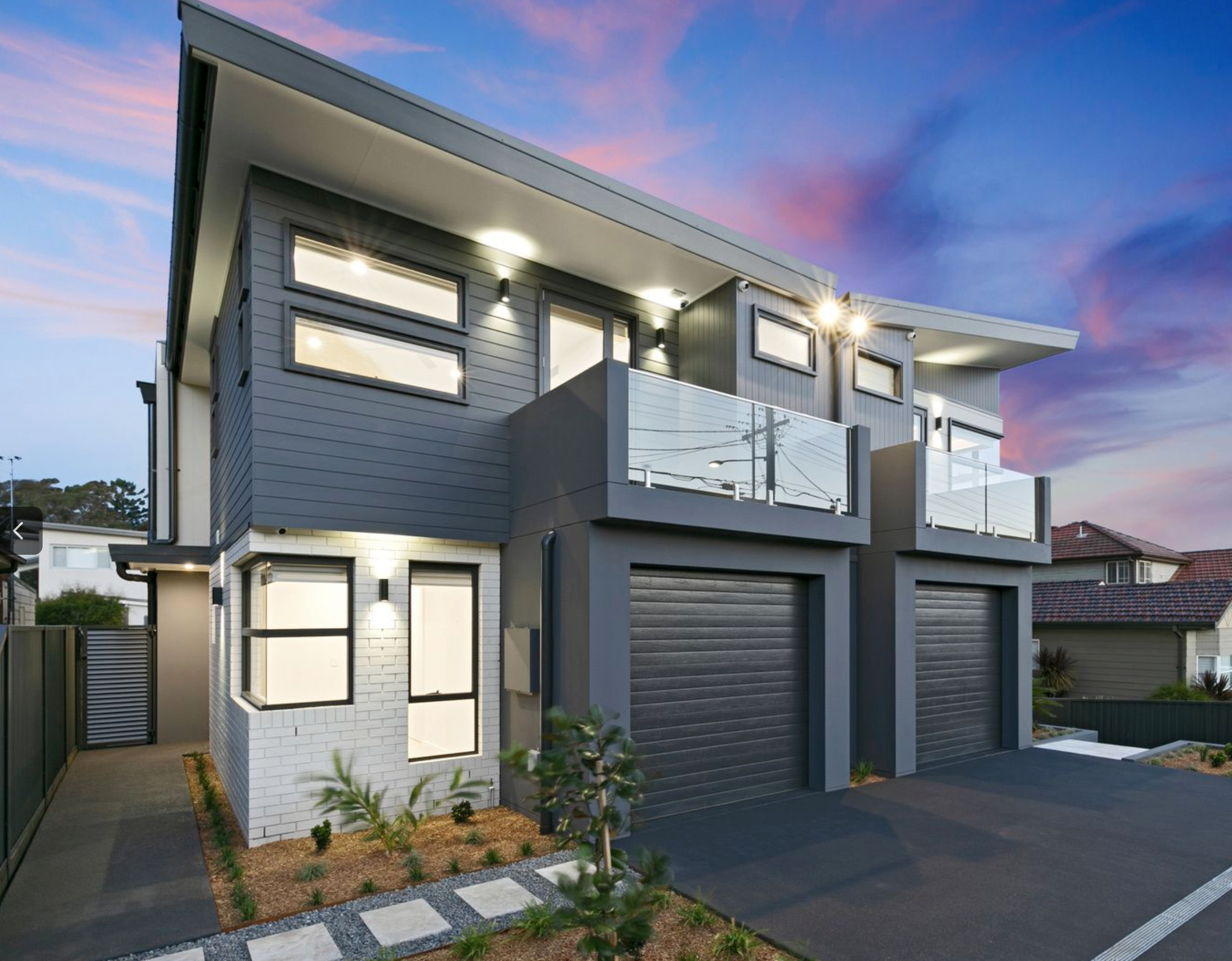 Modern two-story house with gray and white exterior, glass balcony railings, large windows, and a two-car garage at dusk with colorful sky in background.