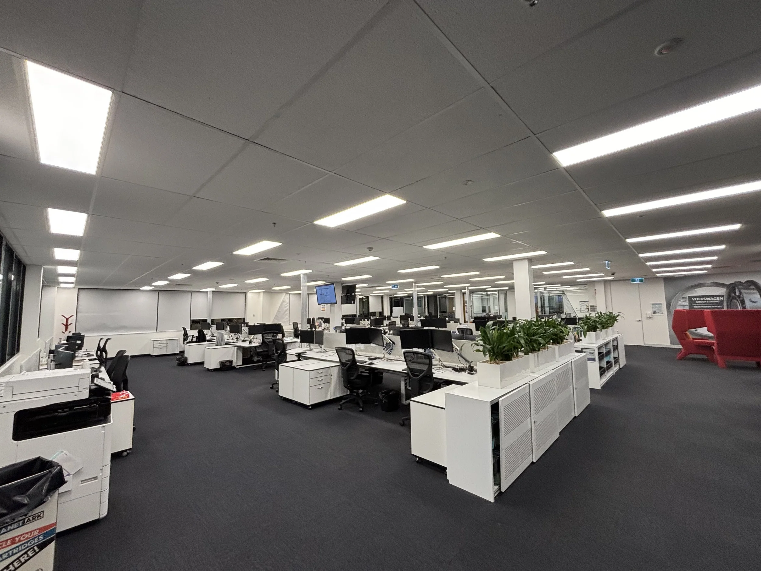 Empty modern office space with white desks and black chairs, plants on some desks, large windows on the left, and a red seating area on the right.