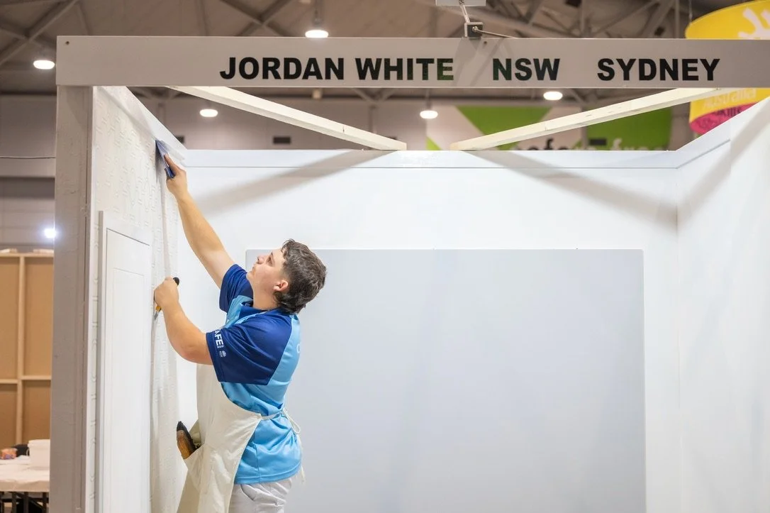 A person installing a white wall panel at an exhibition booth with a sign that reads 'JORDAN WHITE NSW SYDNEY'.