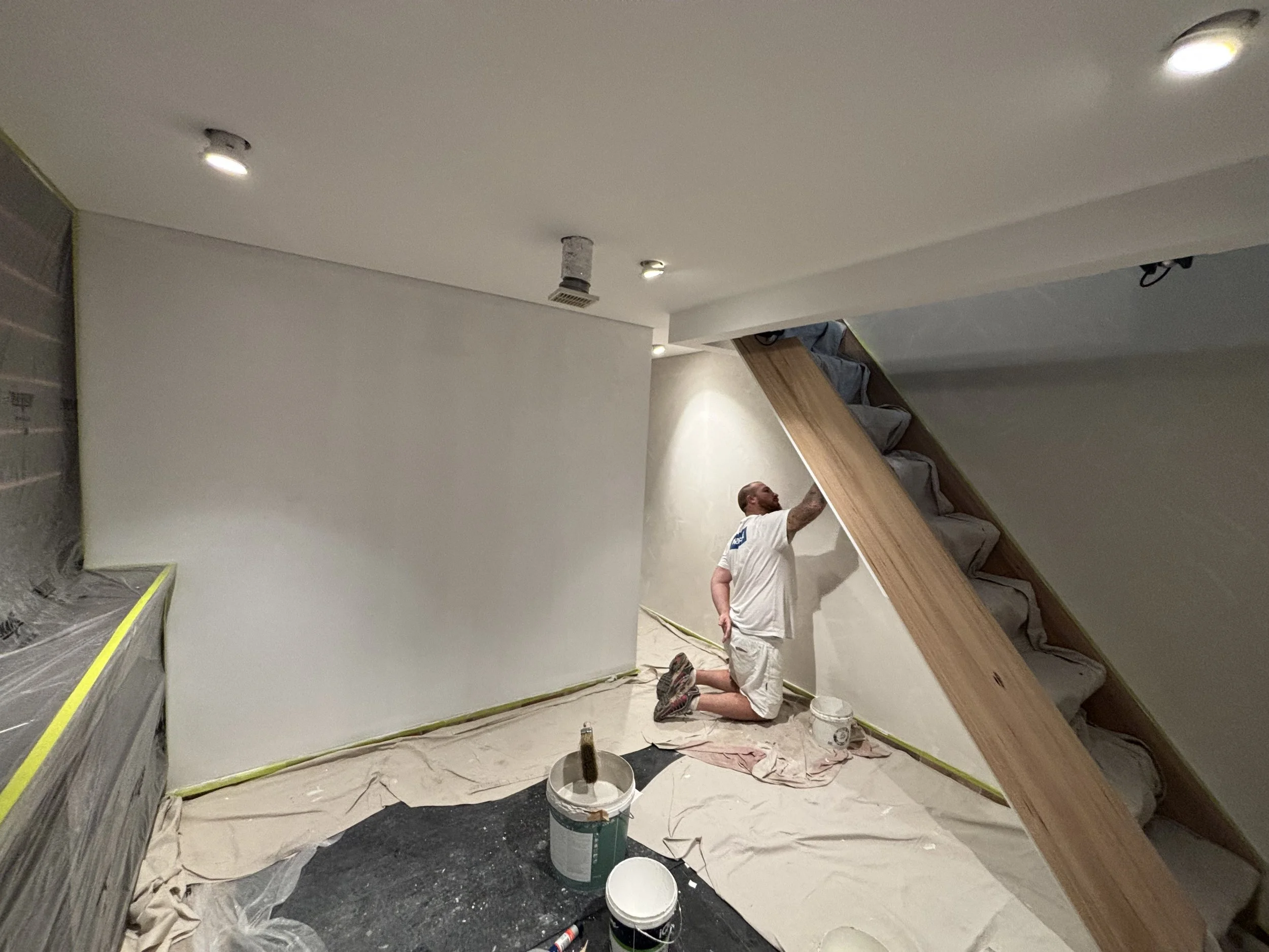 Man kneeling on the floor painting a wall white in a room that appears to be under renovation, with tools and supplies scattered around.