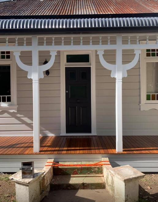 Front porch of a house with white siding, black door, and decorative trim, with stairs under construction in foreground.