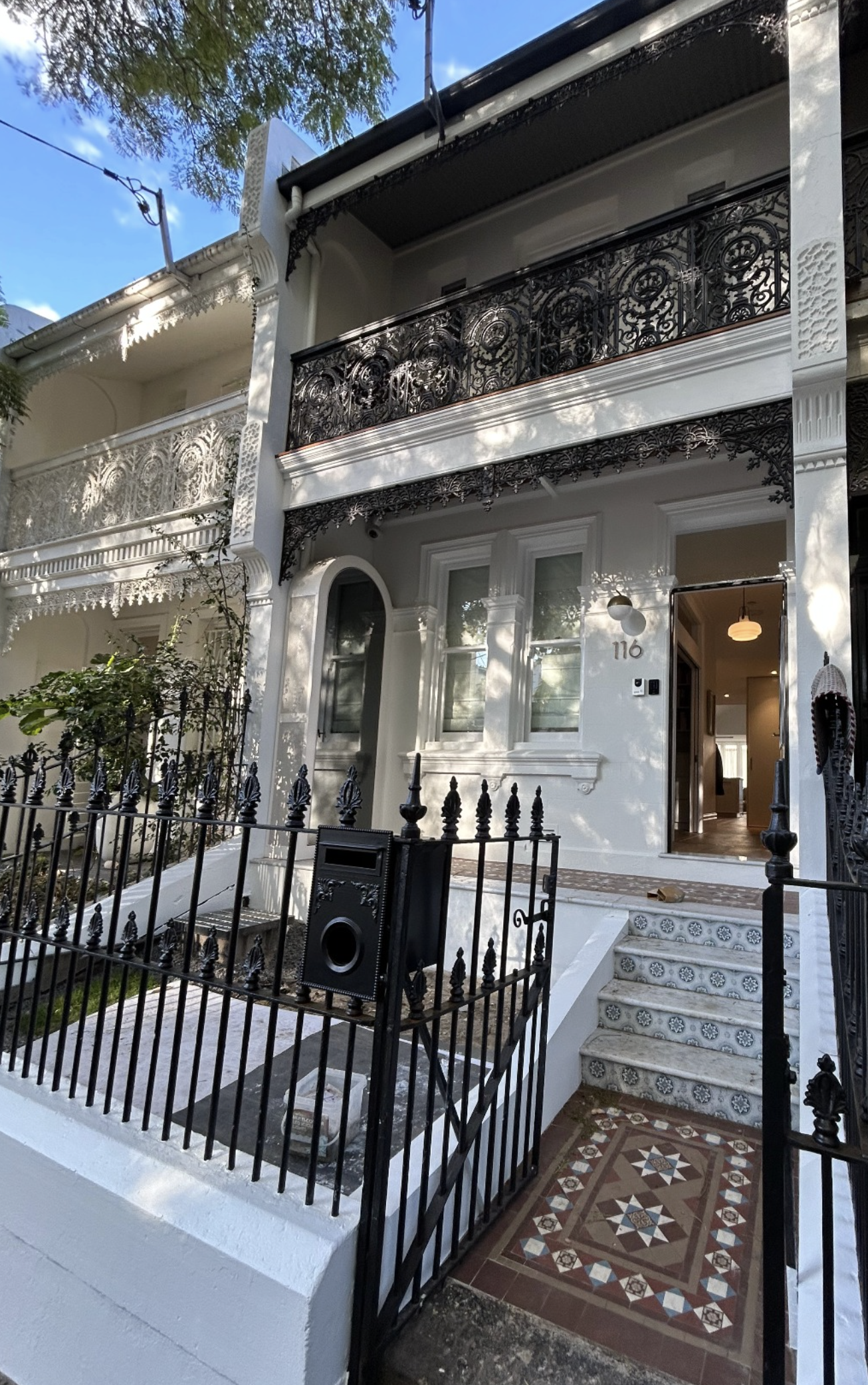 Completed painted Victorian-style house with white walls, black ornate wrought iron balconies, and decorative trim. A black metal fence surrounds the small front yard, which has a patterned tile walkway and a mailbox.
