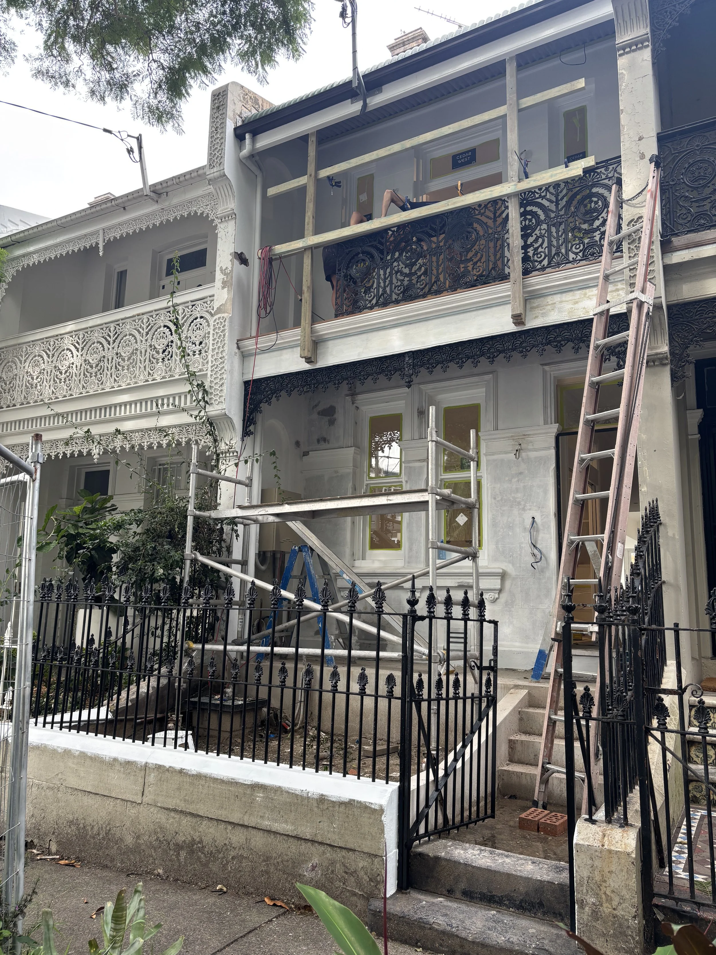 A house undergoing renovation with scaffolding and a ladder, with construction workers working on the upper balcony. Preparation for painting on the exterior of a home in Sydney CBD. Paddington