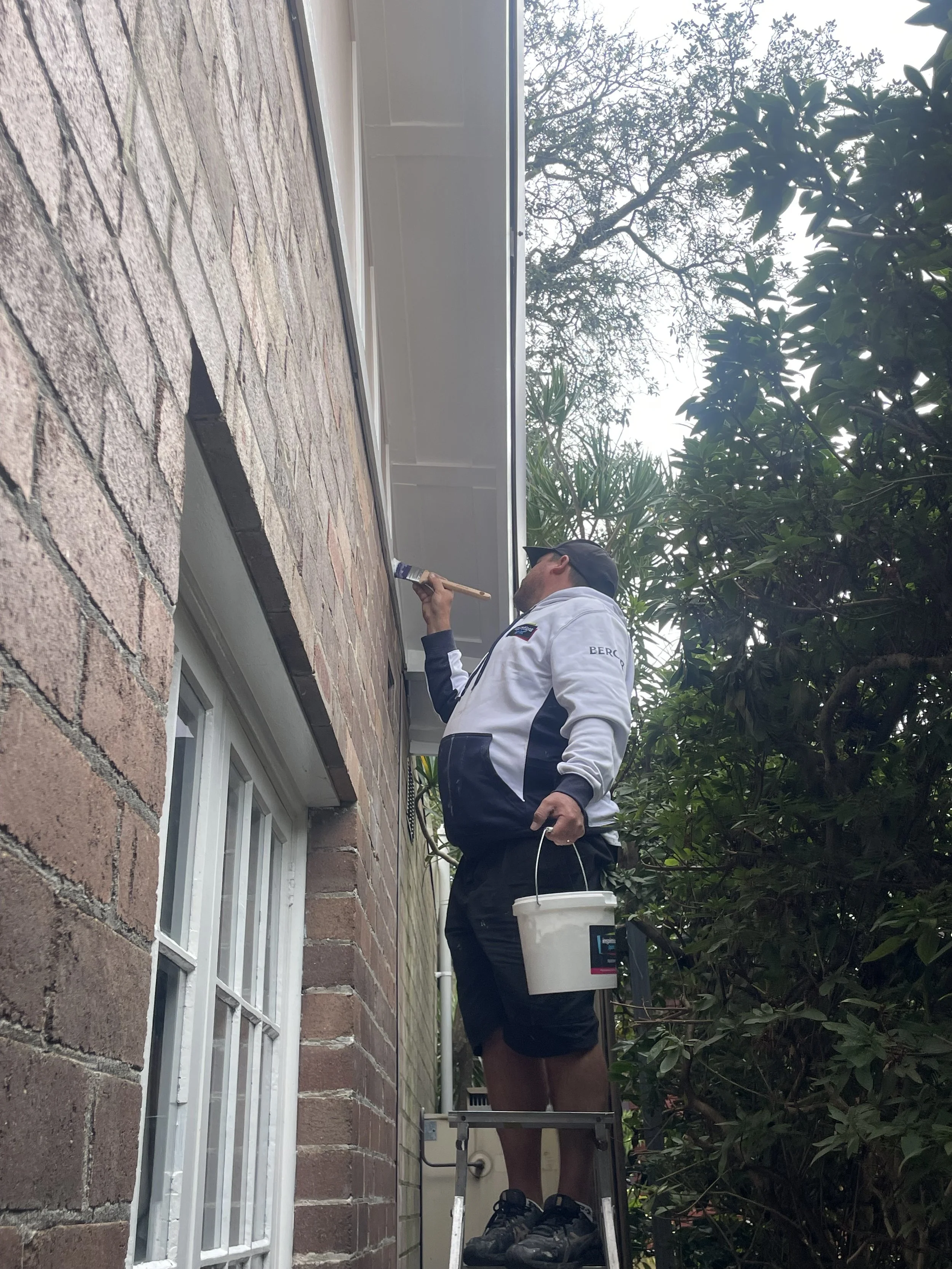 A man painting the exterior wall of a house while standing on a small ladder, holding a bucket and a paintbrush.